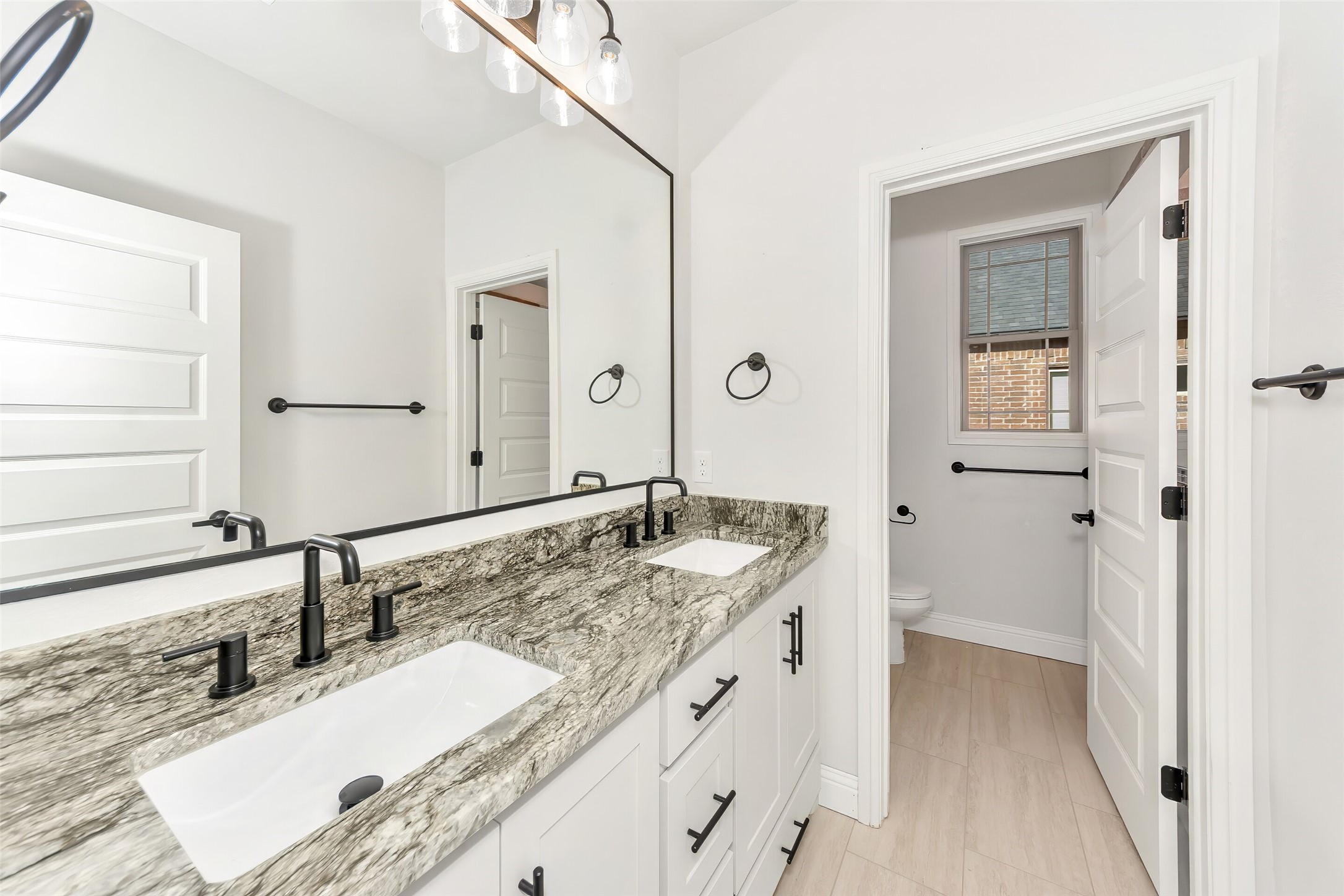 Bathroom with a double-sink granite vanity and an adjoining toilet room with a window.