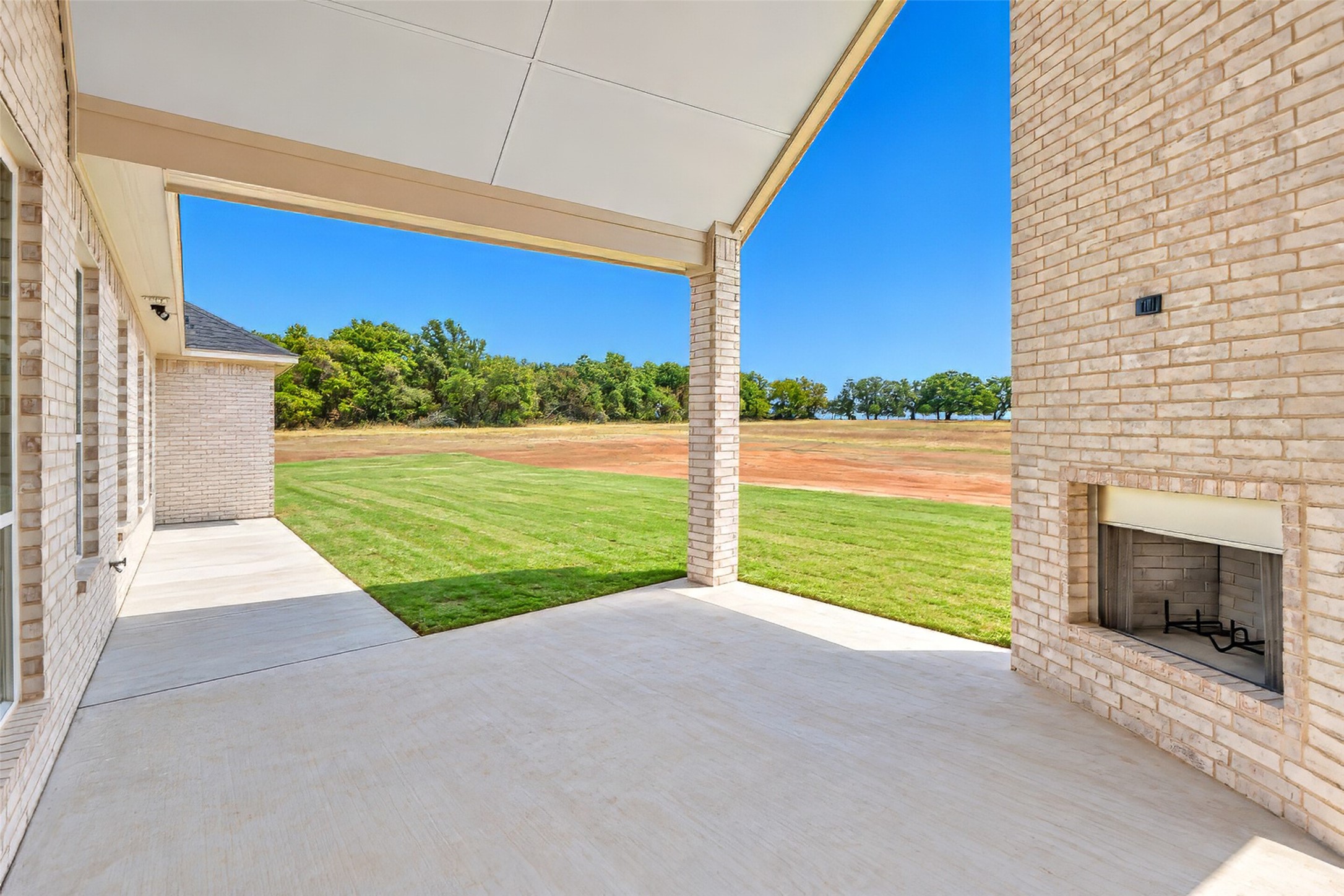 Covered brick patio with an outdoor fireplace overlooking a lawn and open rural land.
