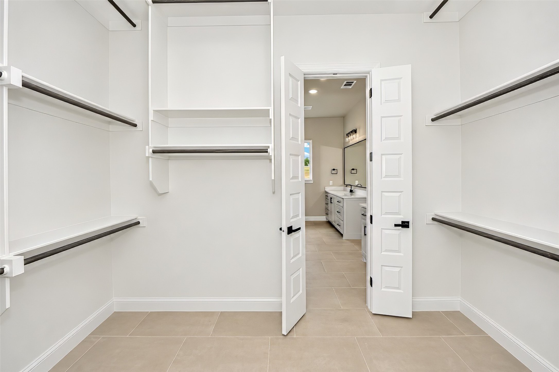 Walk-in closet with built-in shelving and dark hanging rods, opening onto a tiled bathroom vanity.