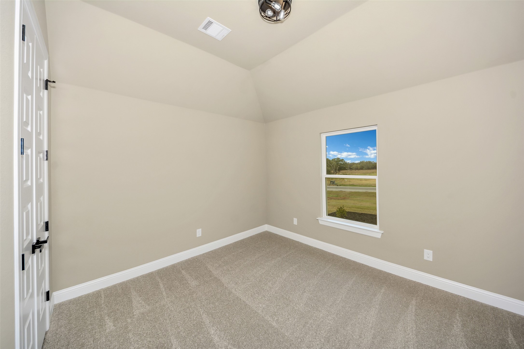 Empty carpeted room with a vaulted ceiling and a window overlooking open land.