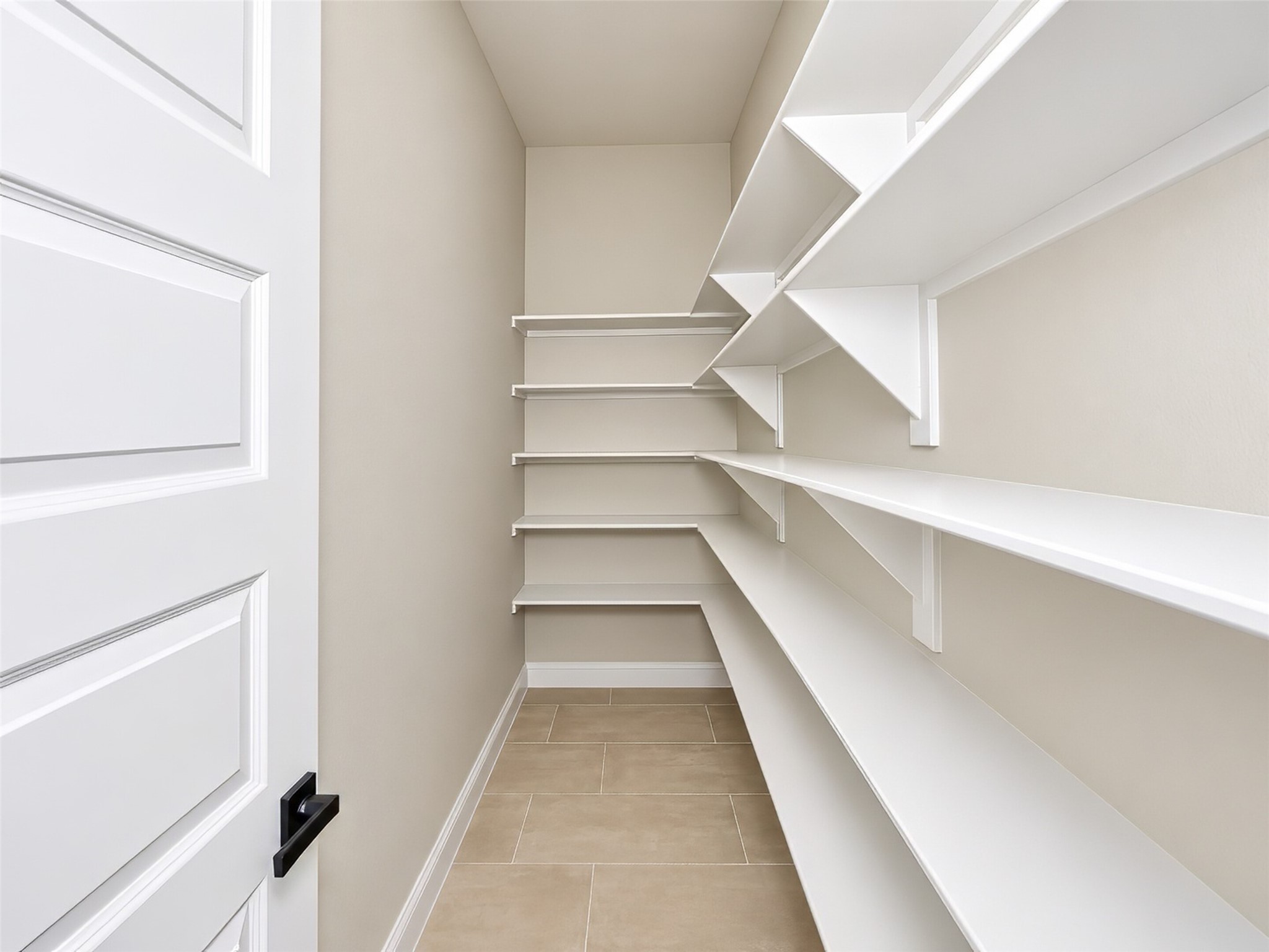 Walk-in pantry with white built-in shelving, beige walls, and tan tile flooring.