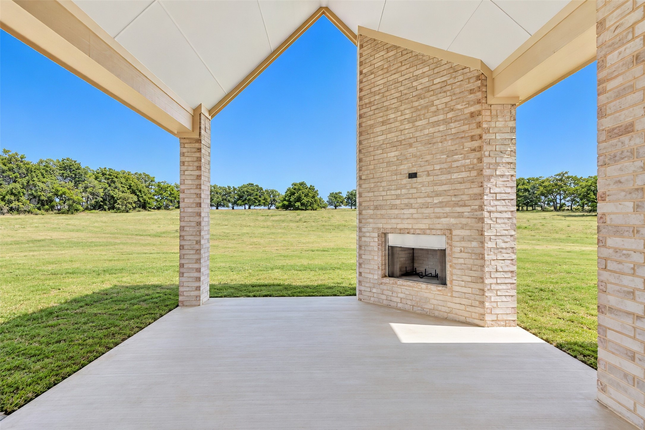 Covered patio with a brick fireplace overlooking a grassy lawn and trees.