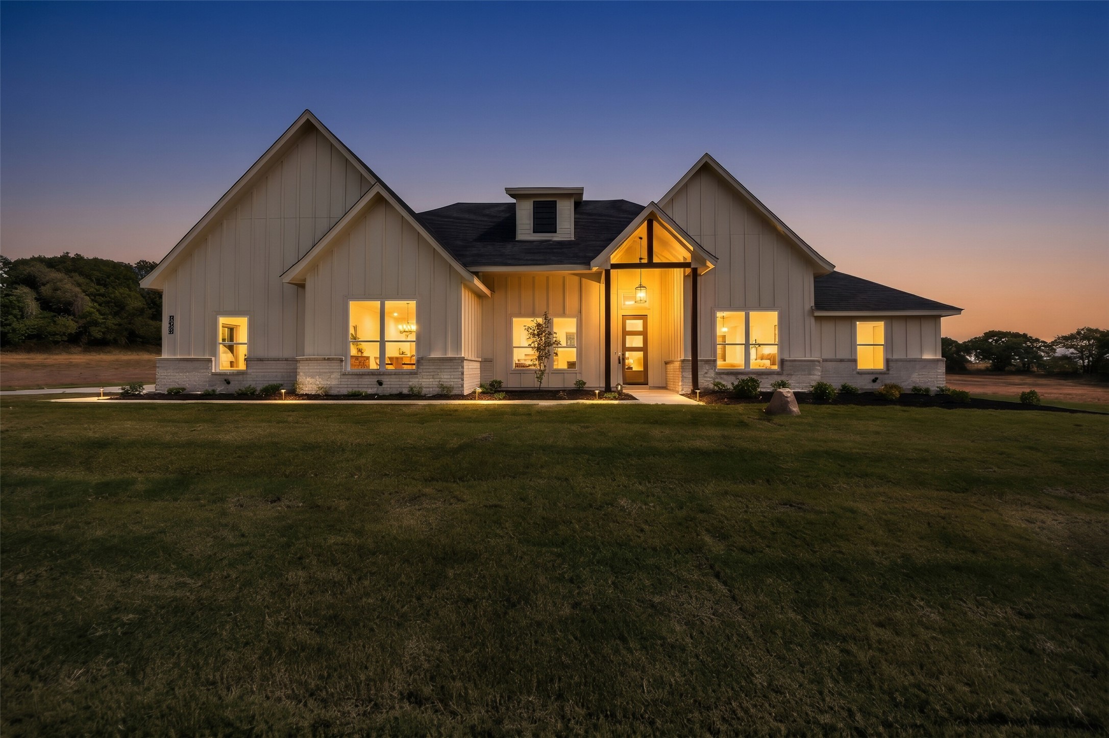 Modern farmhouse exterior at dusk, with steep gables and warmly lit windows overlooking a broad lawn.