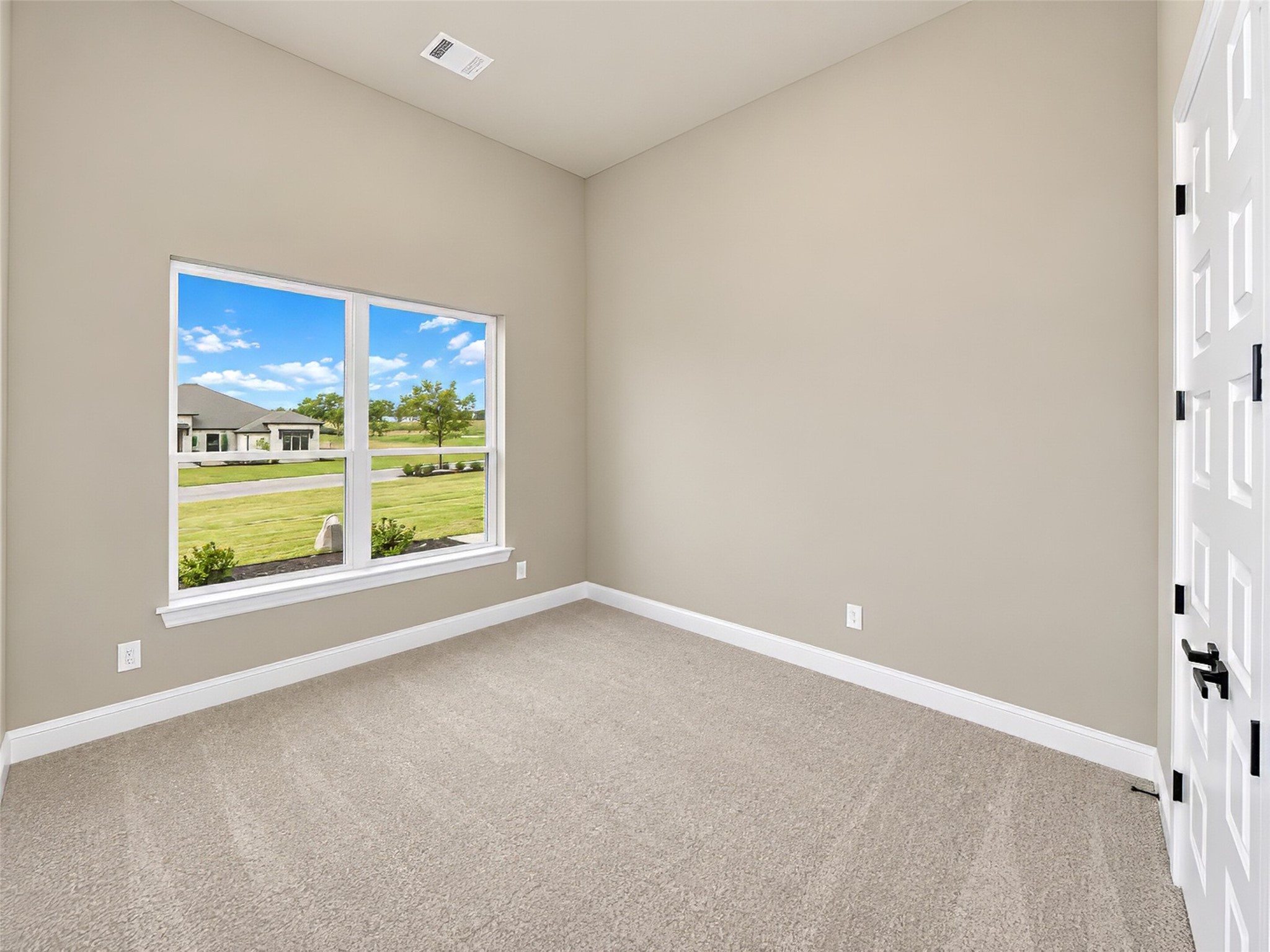 Empty carpeted room with beige walls, a large window overlooking a lawn, and a white paneled door.