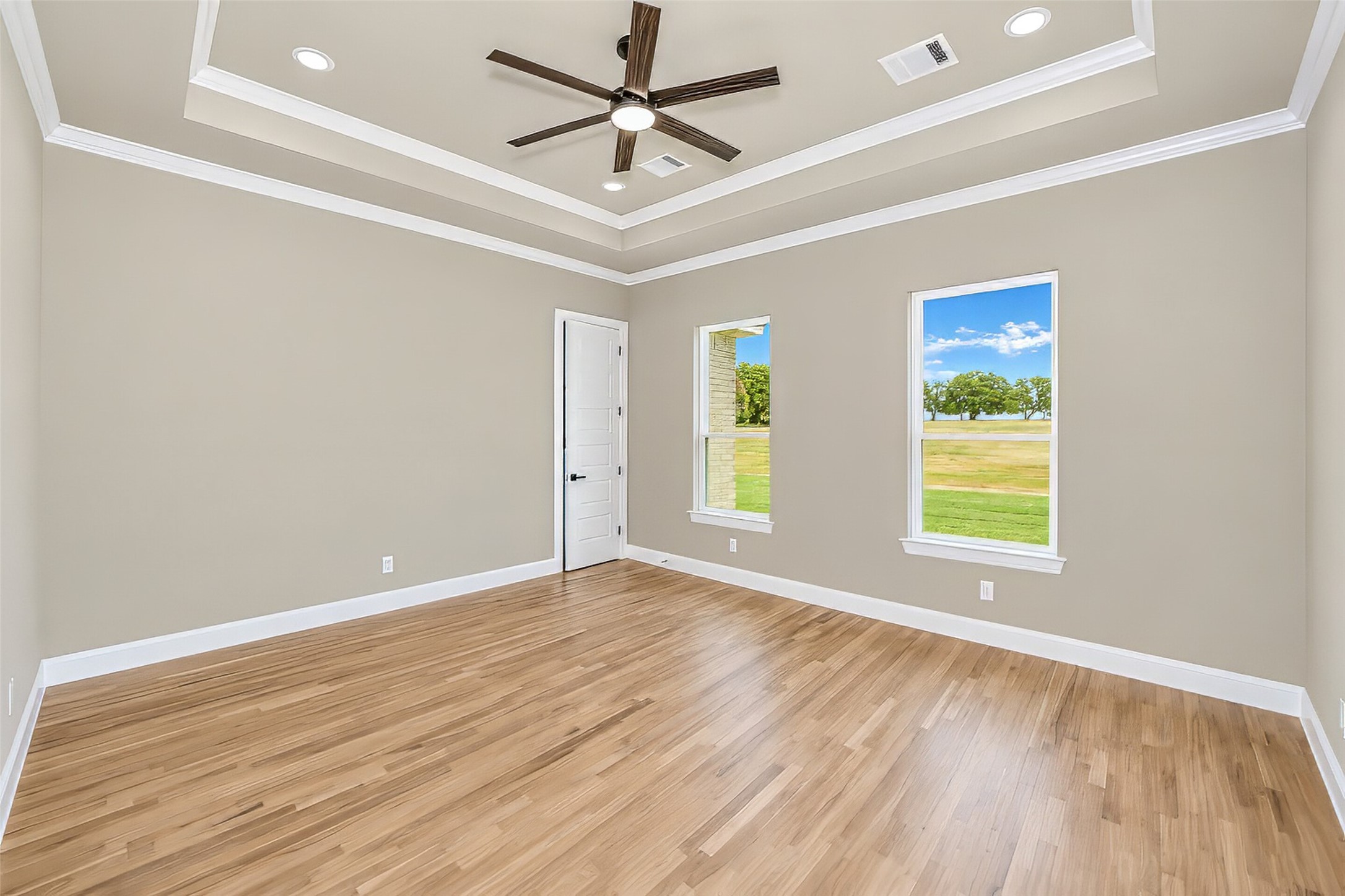 Empty room with warm wood floors, a tray ceiling, ceiling fan, and windows overlooking a green lawn.