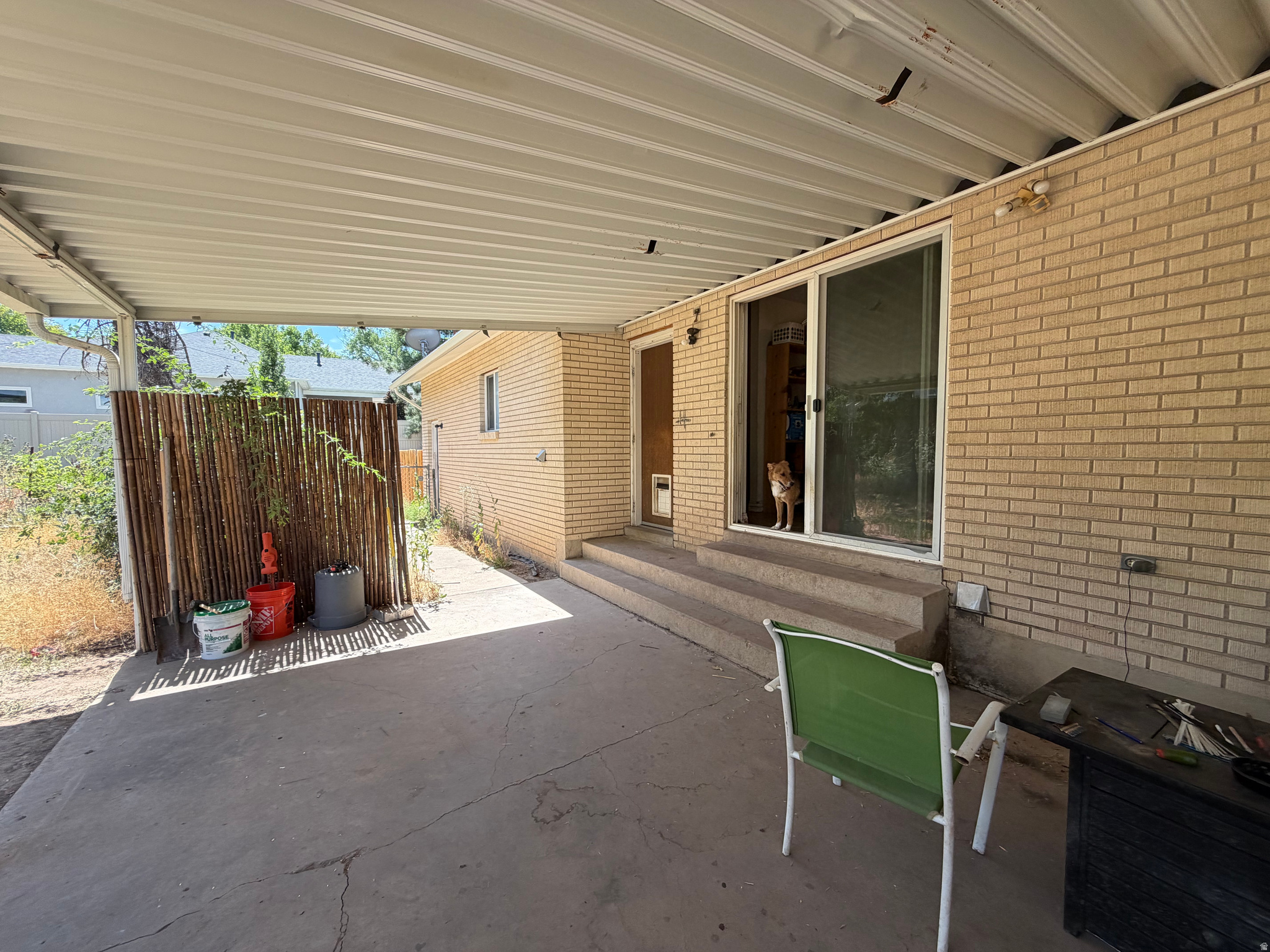 Covered concrete patio with tan brick walls, sliding doors, a wooden privacy screen, and a dog in the doorway.