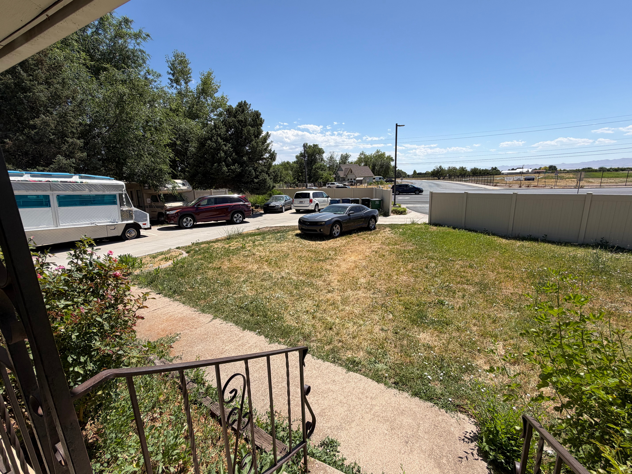 Front yard and walkway lead to a driveway with parked vehicles beside a tall fence.