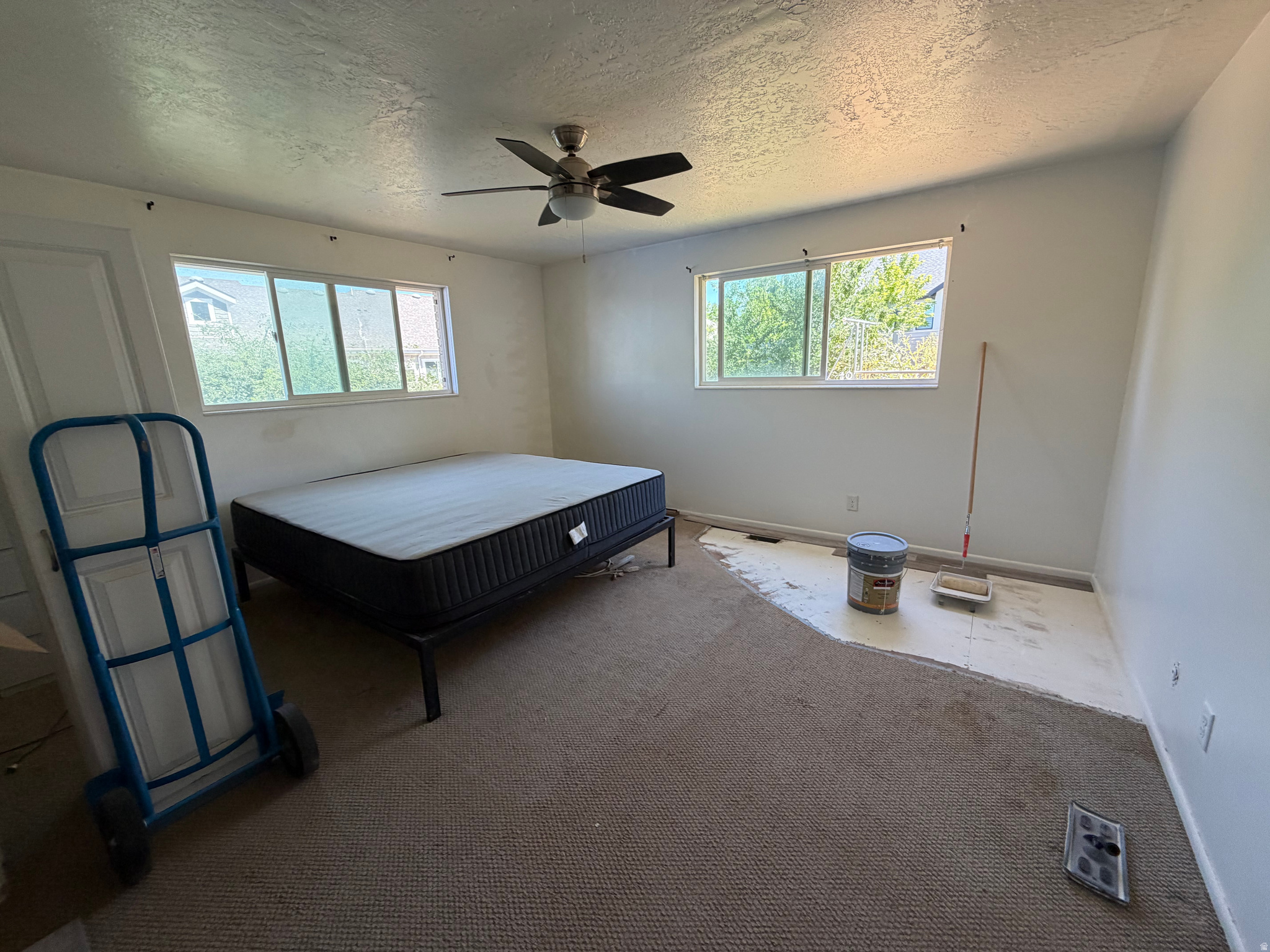 Unfinished bedroom with a mattress, two large windows, and exposed flooring beside the carpet.