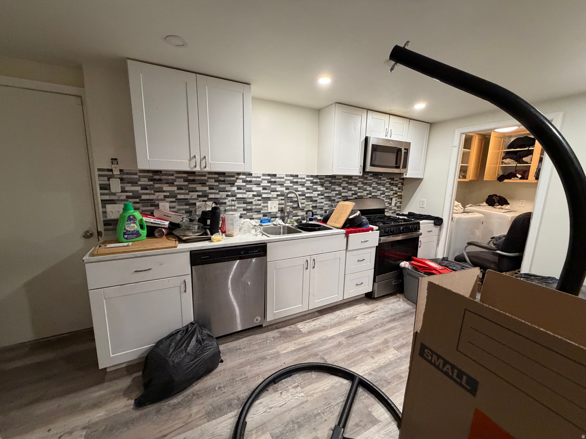 Kitchen with white cabinets, mosaic tile backsplash, stainless appliances, and cluttered counters.