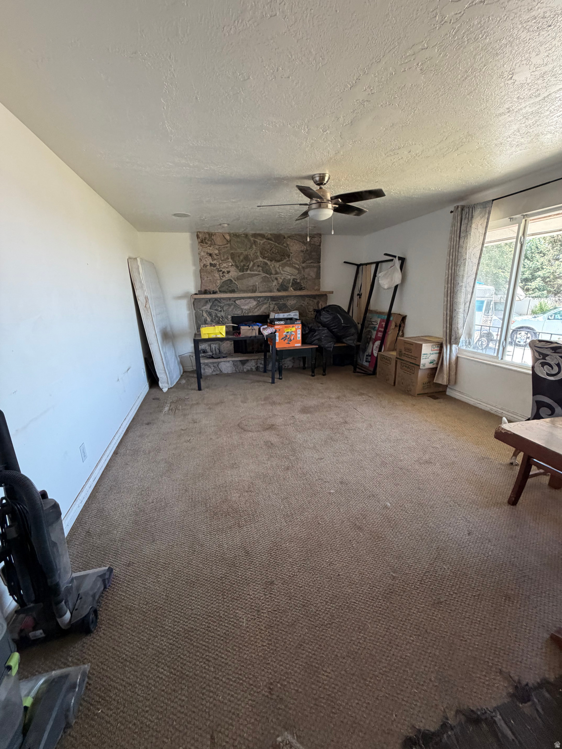 Carpeted living room with a stone fireplace, ceiling fan, and boxes by the window.