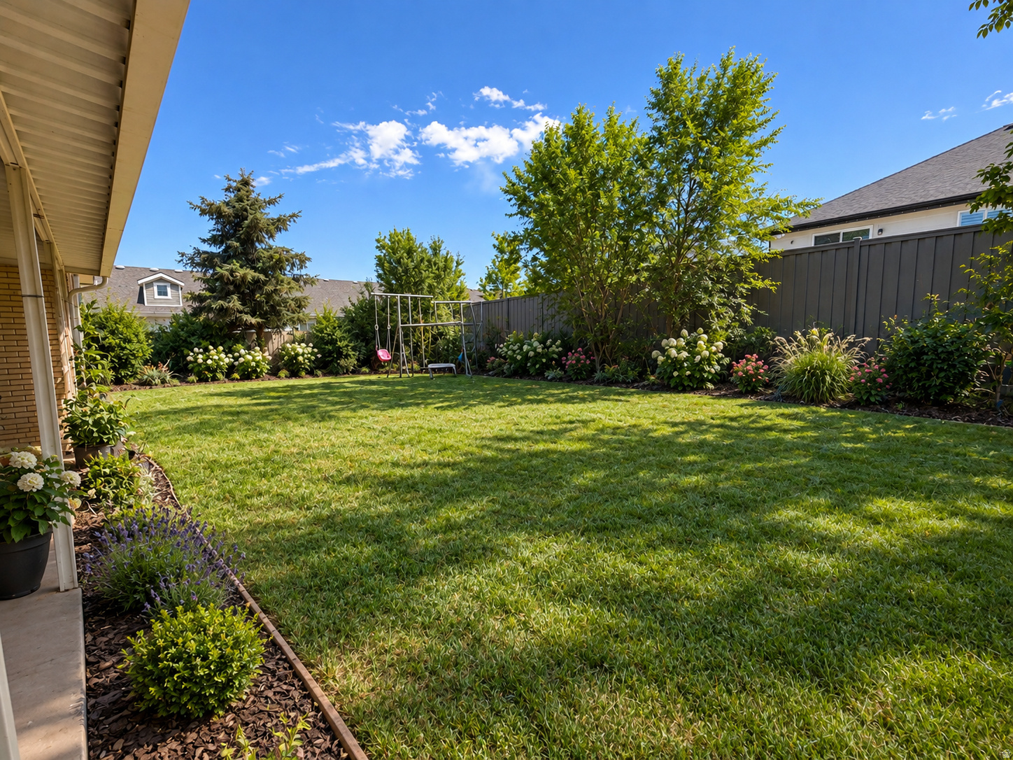 Backyard lawn bordered by flower beds and privacy fencing, with a swing set and covered patio.