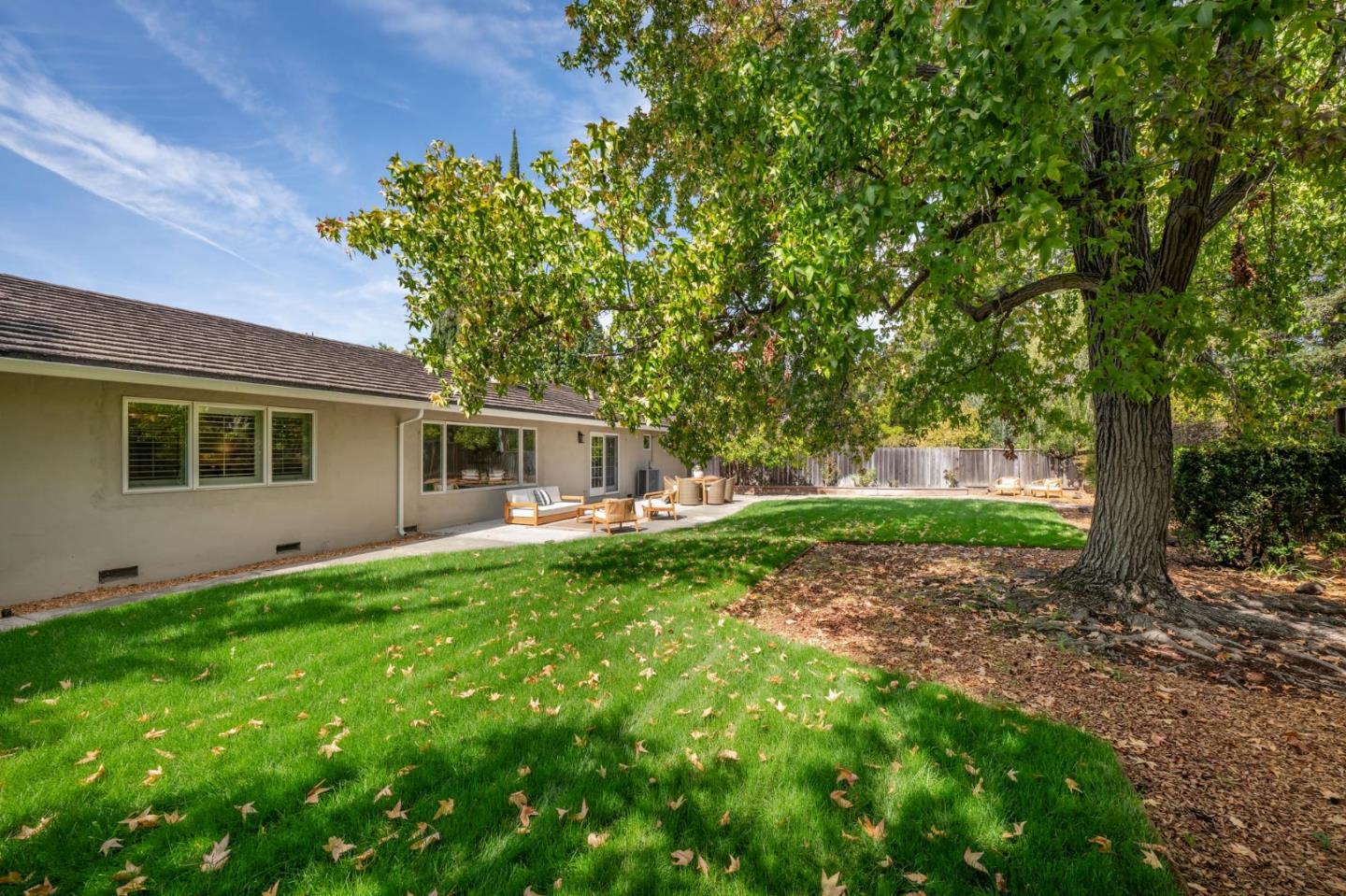 Home’s rear yard with a broad lawn, mature shade tree, and patio seating.