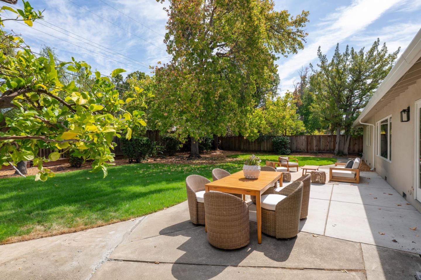 Backyard patio with a wicker dining set beside a lawn and mature trees.