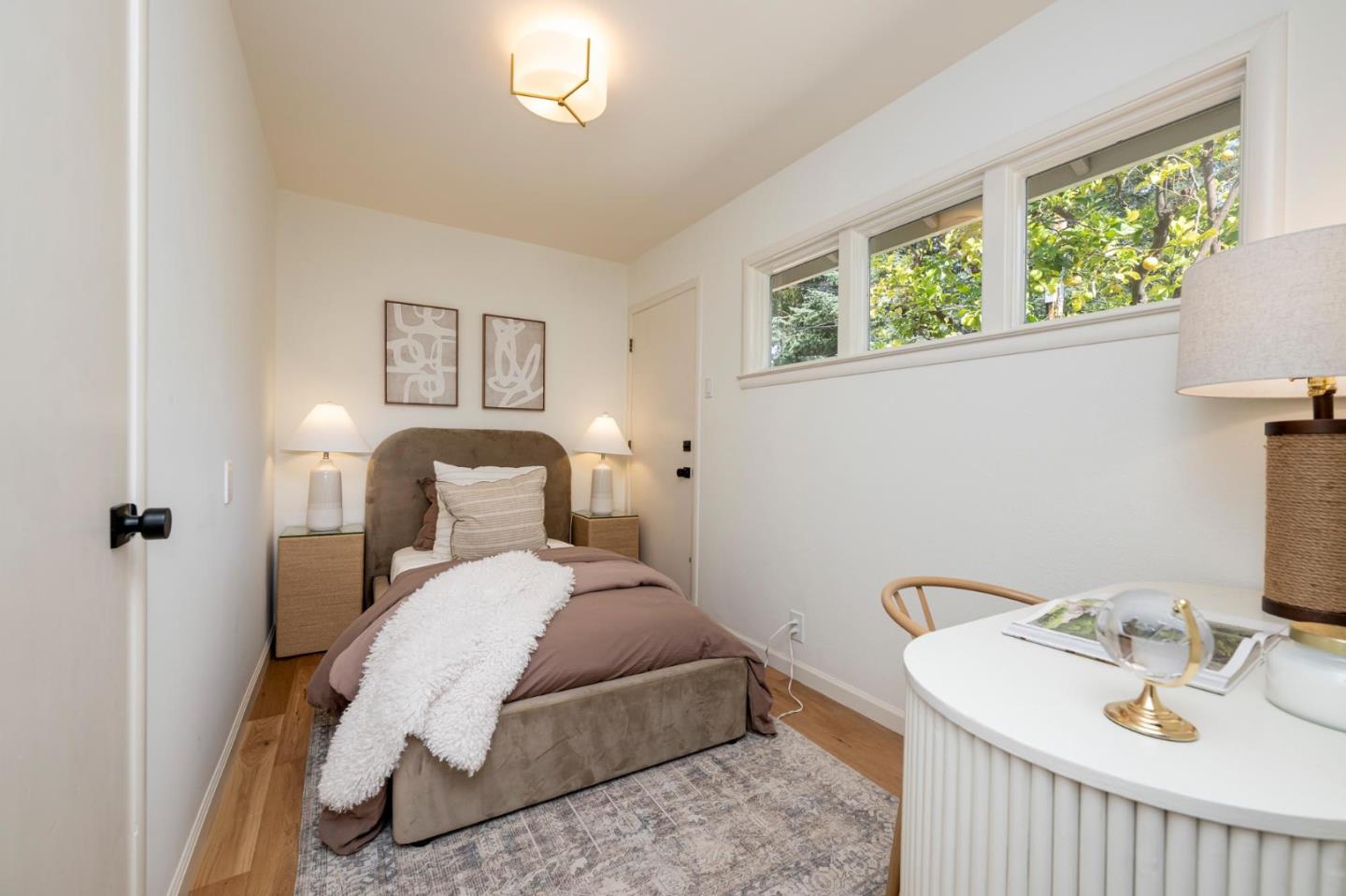 Staged bedroom with a taupe bed, textured white throw, and long windows overlooking greenery.