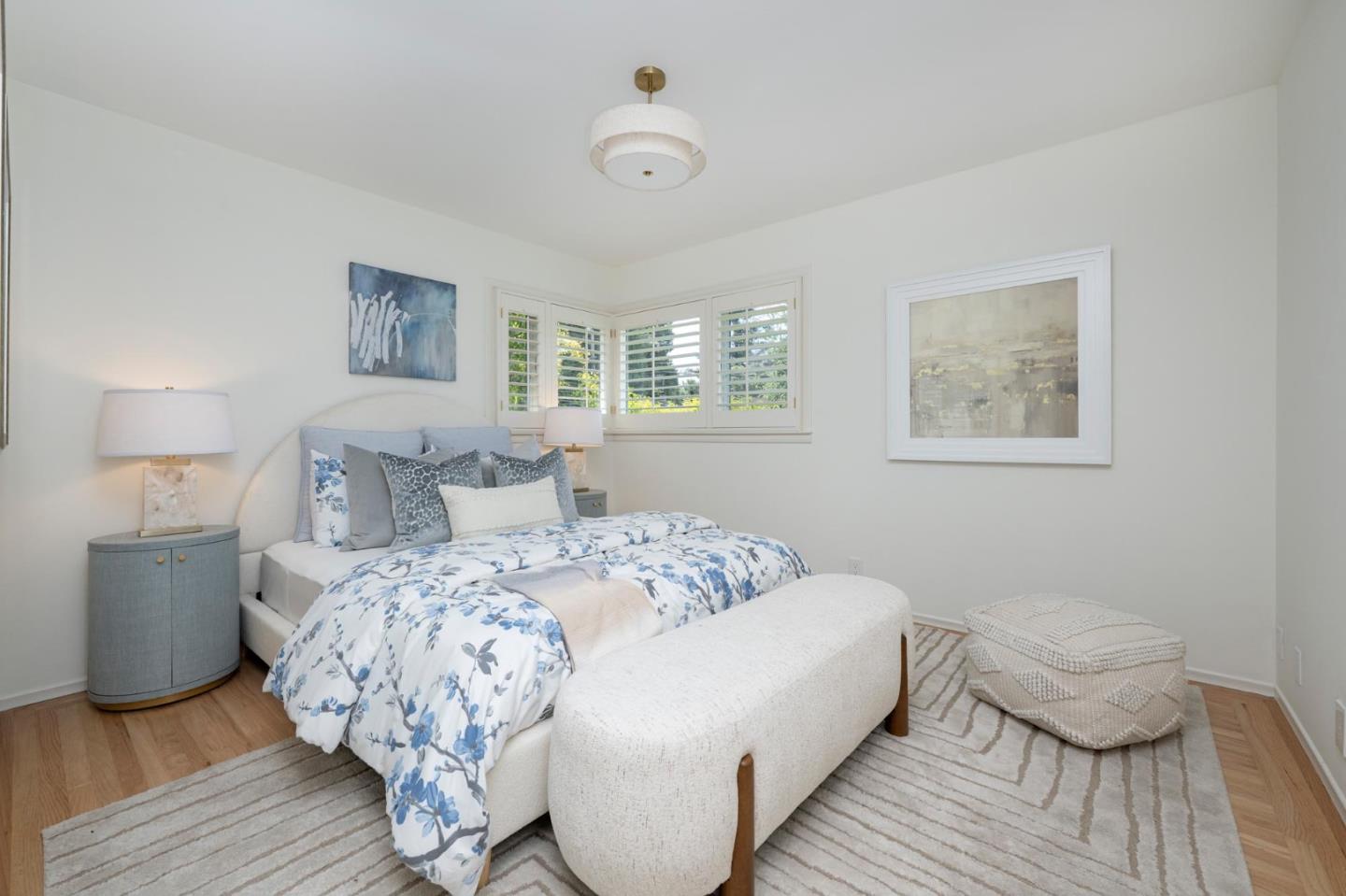 Bright bedroom with blue floral bedding, a curved bench, and plantation-shuttered corner windows.