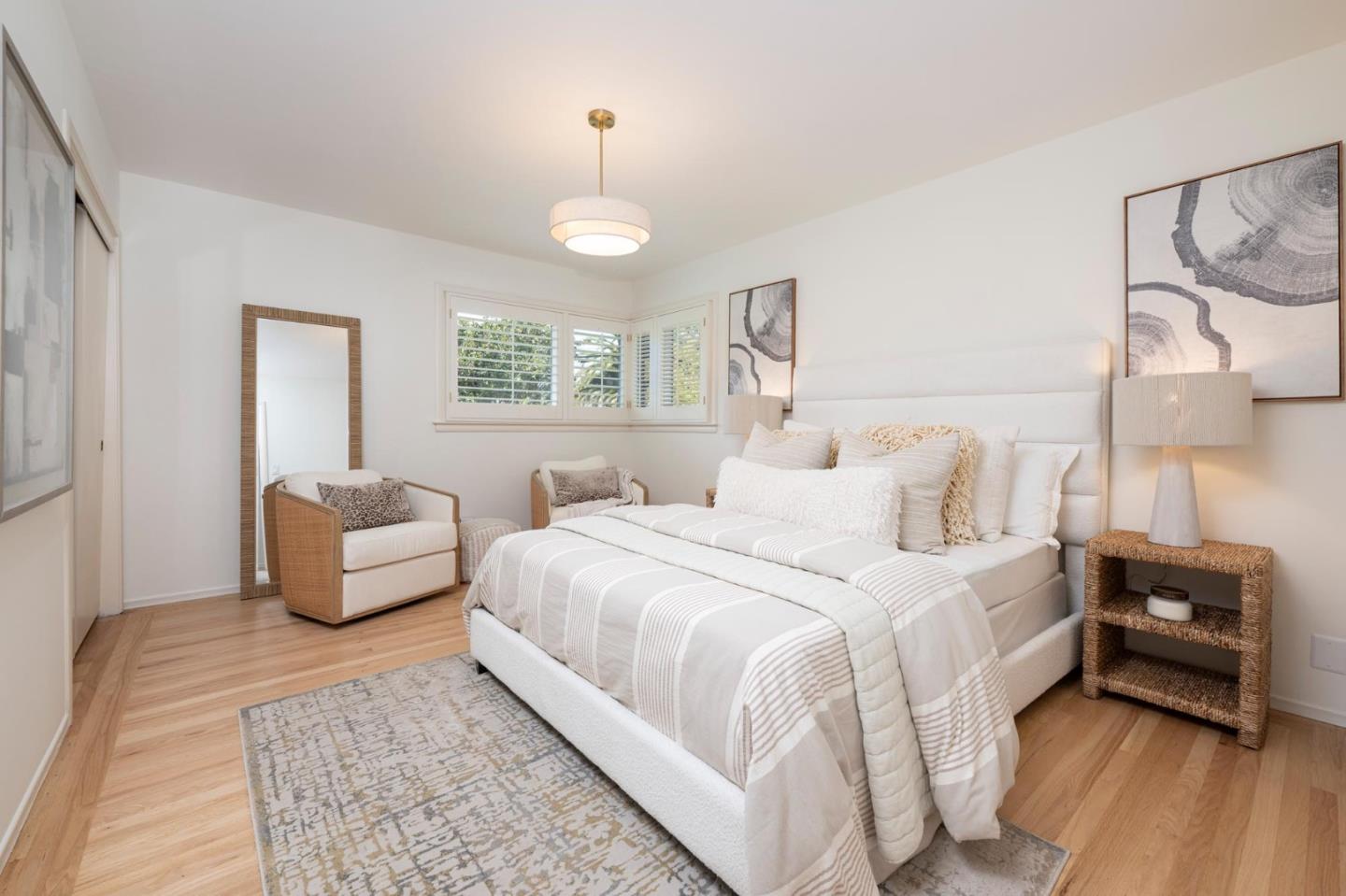 Staged bedroom with a white upholstered bed, neutral bedding, and two armchairs beside shuttered windows.