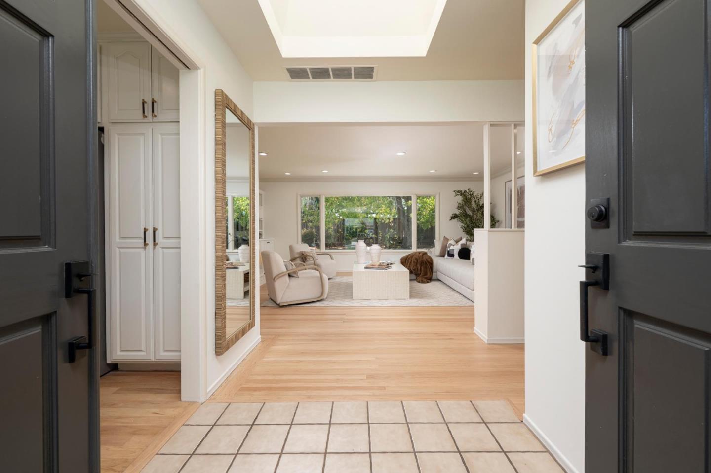 Bright living room beyond a tiled entry, with white seating, hardwood floors, and large garden-facing windows.