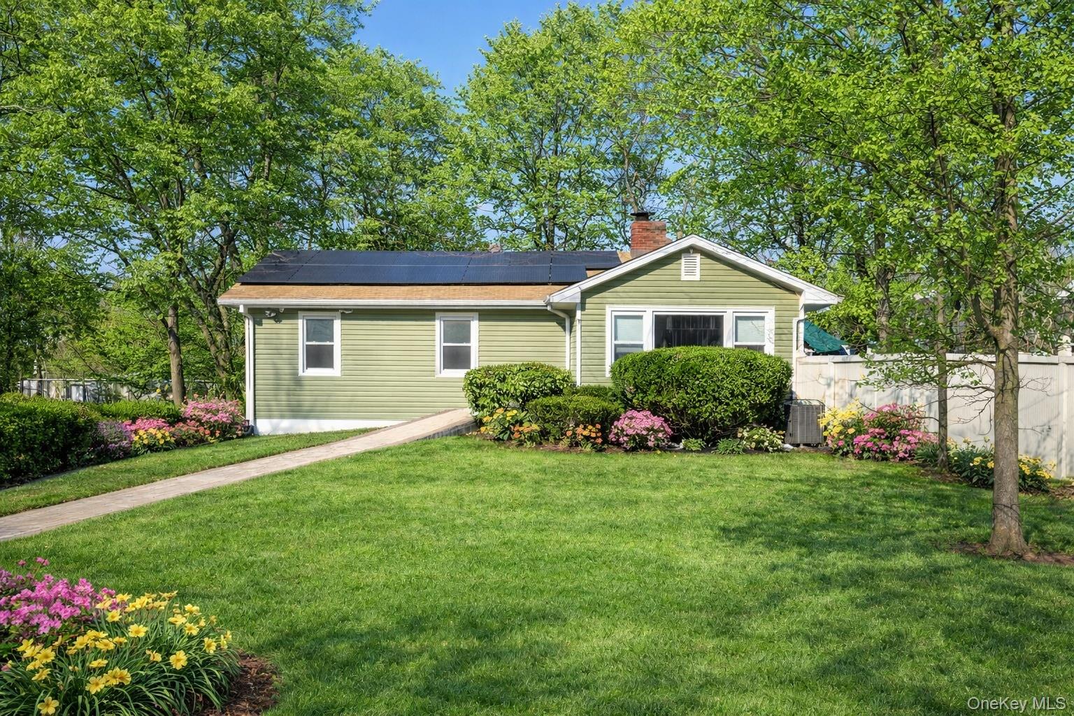 Light green single-story house with rooftop solar panels, a curved walkway, and colorful flower beds.