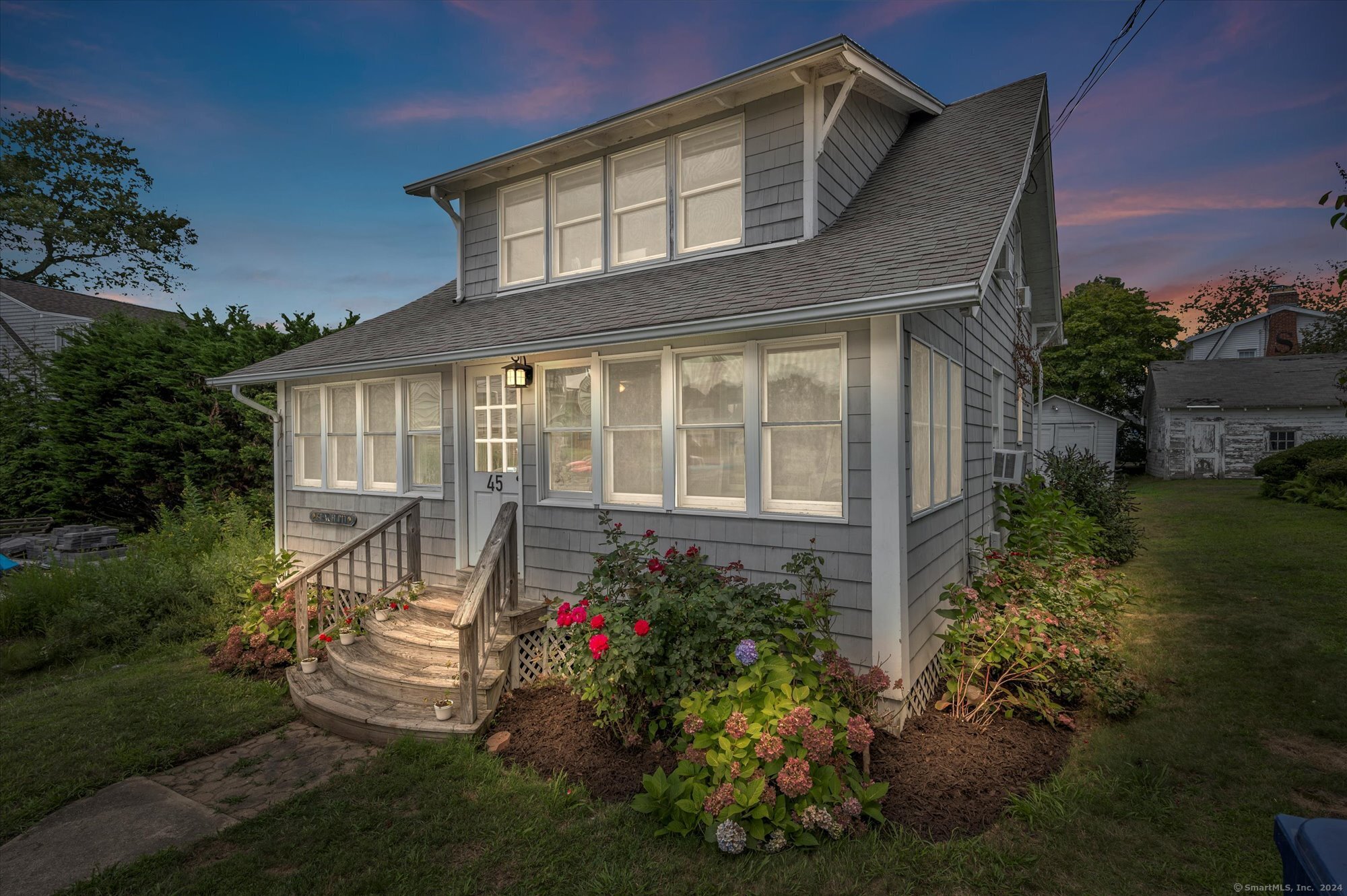 Gray shingle-sided house with an upper dormer, curved wooden steps, and colorful flower beds at dusk.