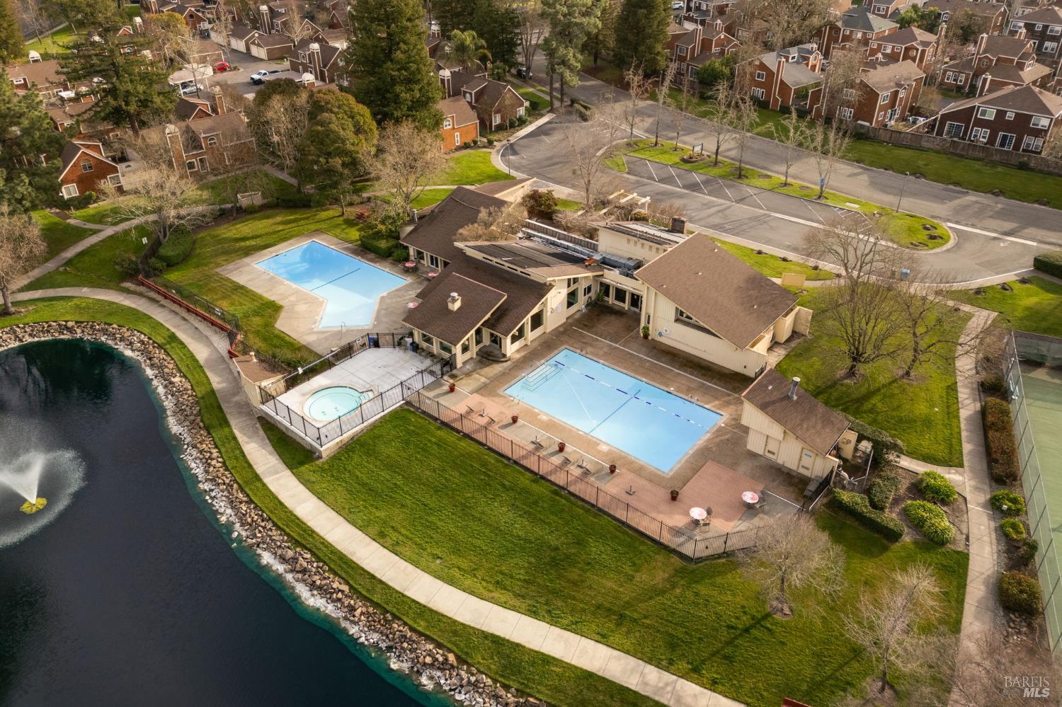 Aerial view of a community clubhouse and pools beside a lake, with nearby homes and a tennis court.