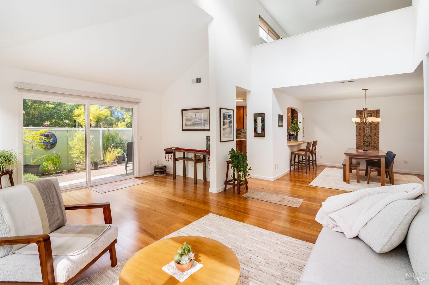 Open-plan living and dining room with vaulted ceilings, hardwood floors, and sliding doors to a planted patio.