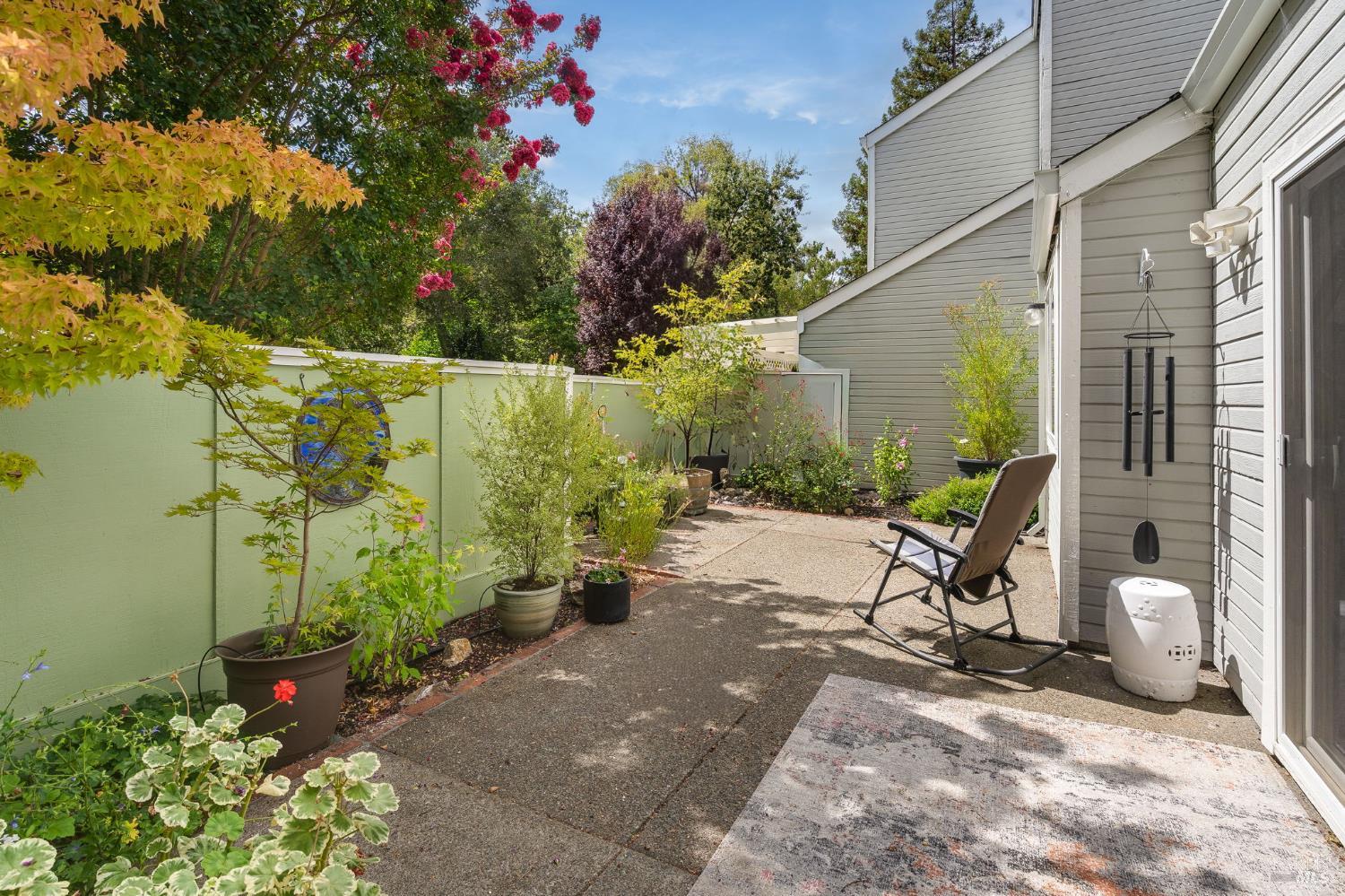 Backyard patio with potted plants, a pale green fence, and a rocking chair beside the house.