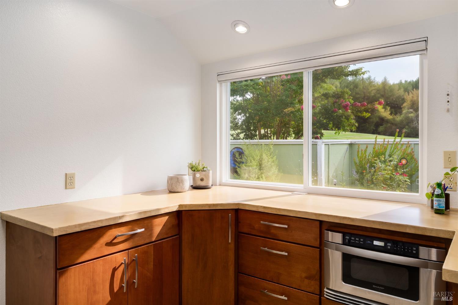 Kitchen with warm wood cabinets and a large window overlooking flowering shrubs.