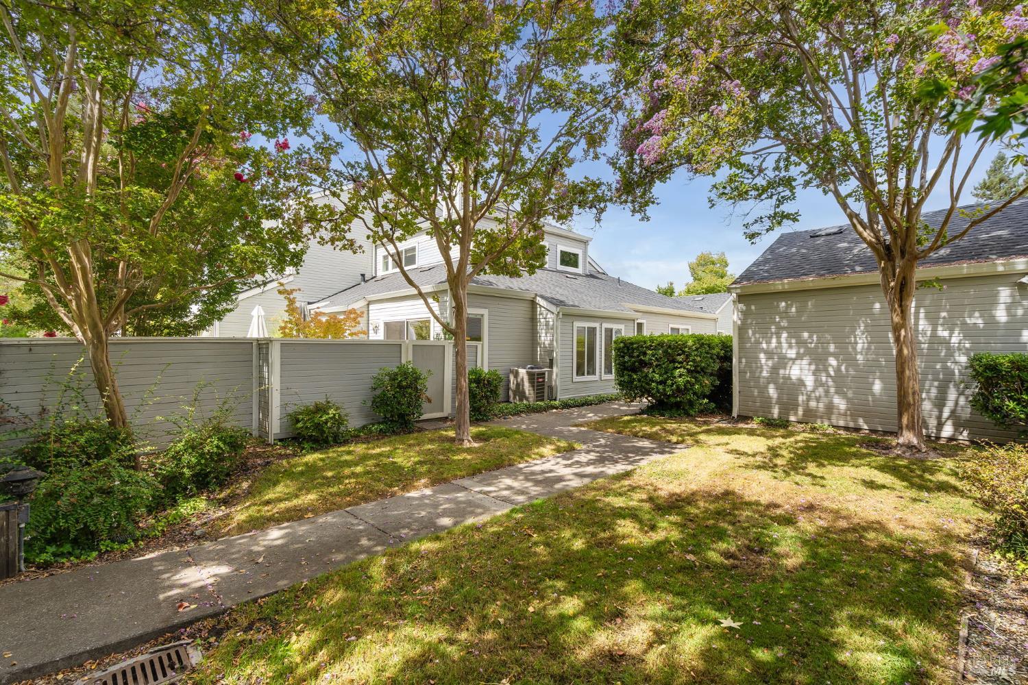 Gray house and detached outbuilding behind a paved path, privacy fence, and flowering trees.