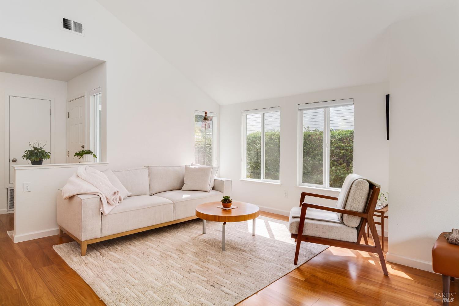 Bright living room with vaulted ceiling, cream sofa, wood-framed armchair, and large windows overlooking hedges.