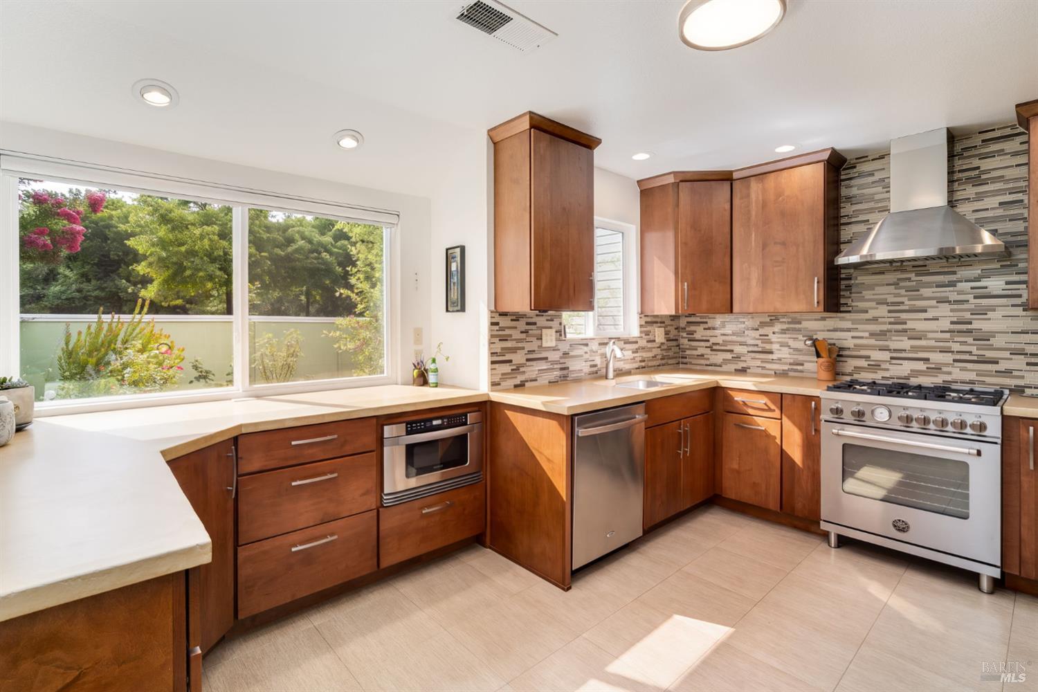 Modern kitchen with warm wood cabinets, stainless steel appliances, and a large window overlooking greenery.