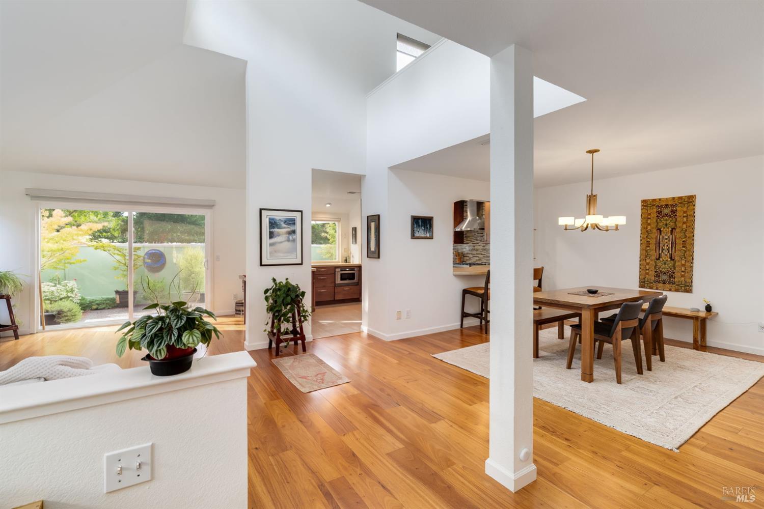 Open-plan living and dining area with tall ceilings, warm wood floors, and garden-facing sliding glass doors.