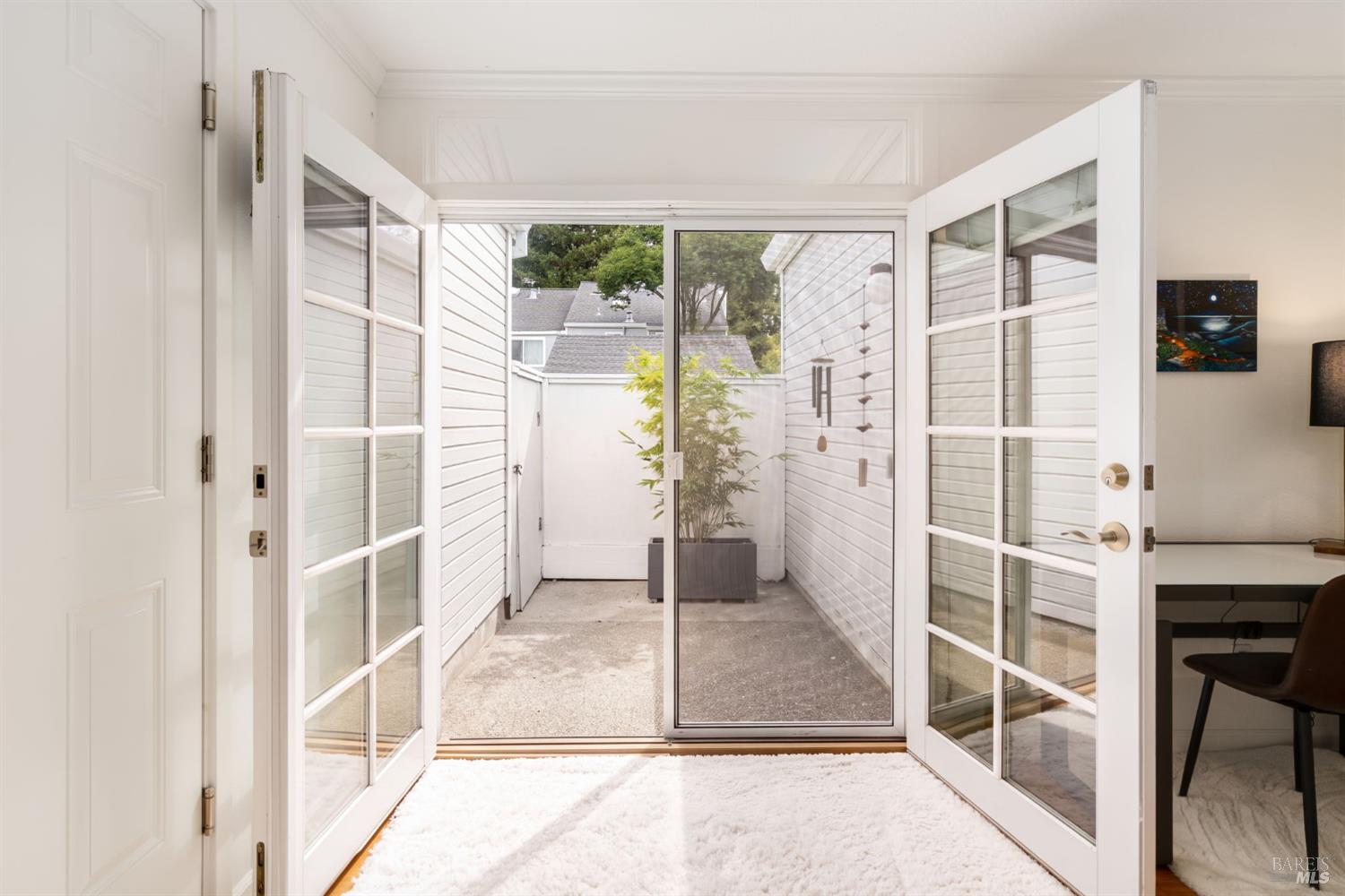 Open French doors lead from a bright room to a small enclosed patio with a potted plant.