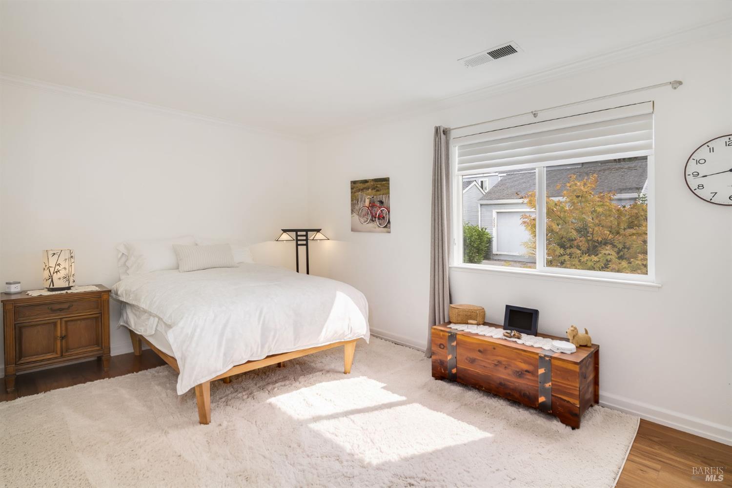 Furnished bedroom with a white bed, cream rug, wooden chests, and a wide sunlit window.