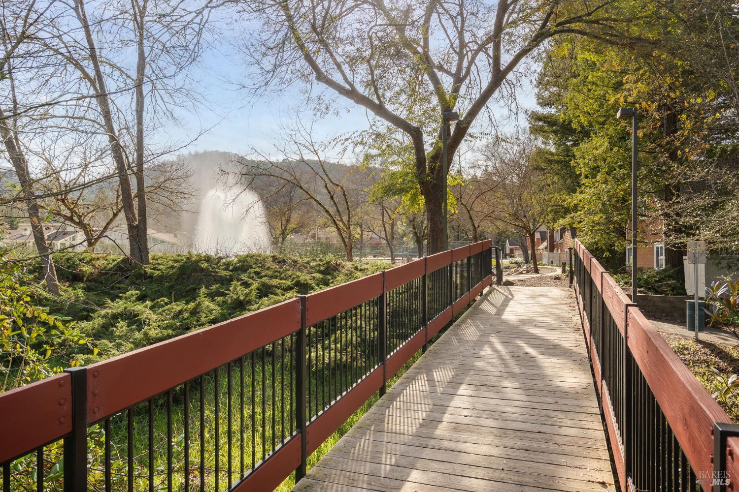 Pedestrian bridge with red-brown railings leads through wooded grounds toward a fountain.
