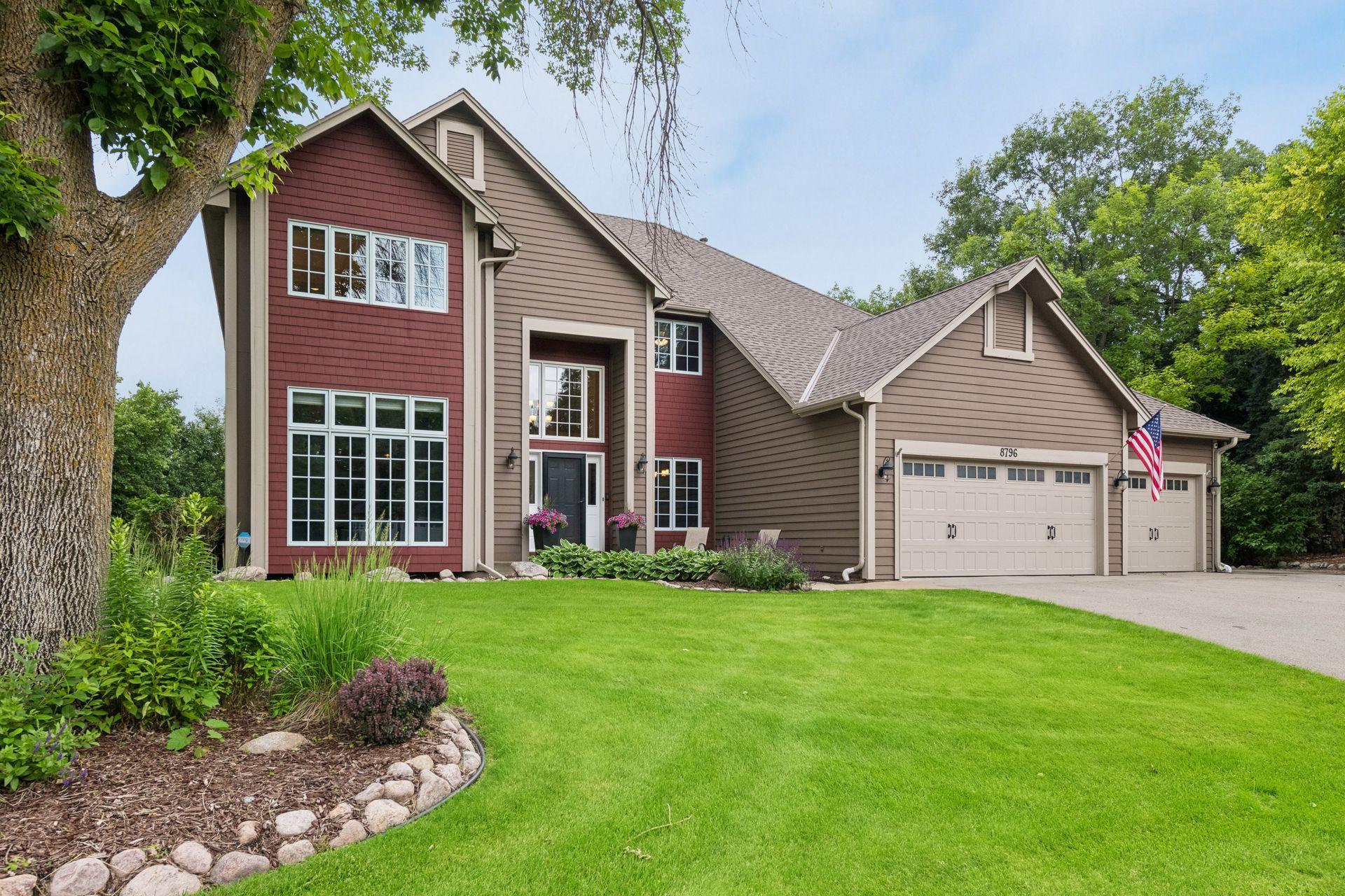 Two-story red and taupe house with a three-car garage, landscaped yard, and American flag.