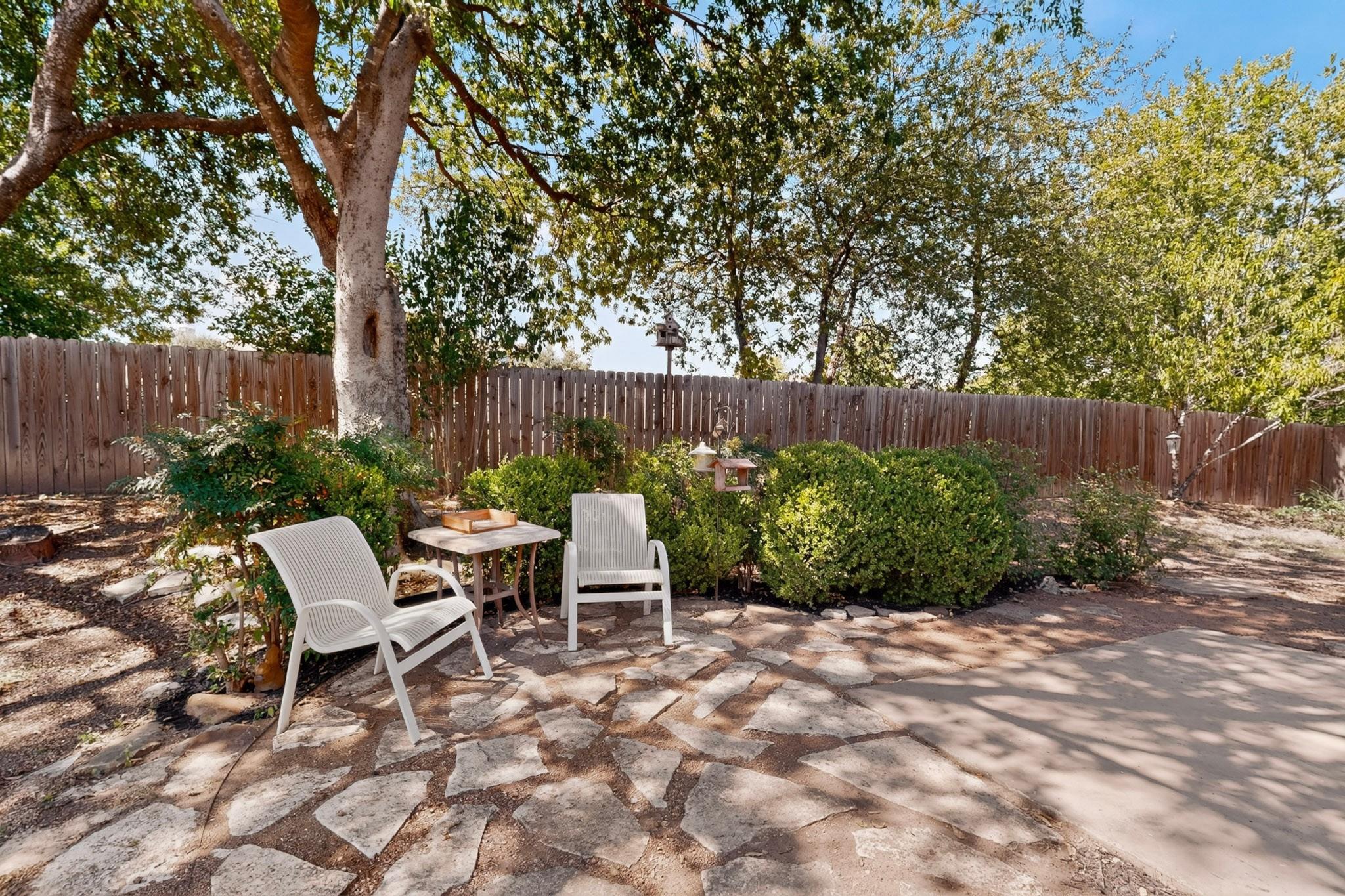 Backyard stone patio with white chairs and table beneath mature trees, bordered by shrubs and a wooden fence.