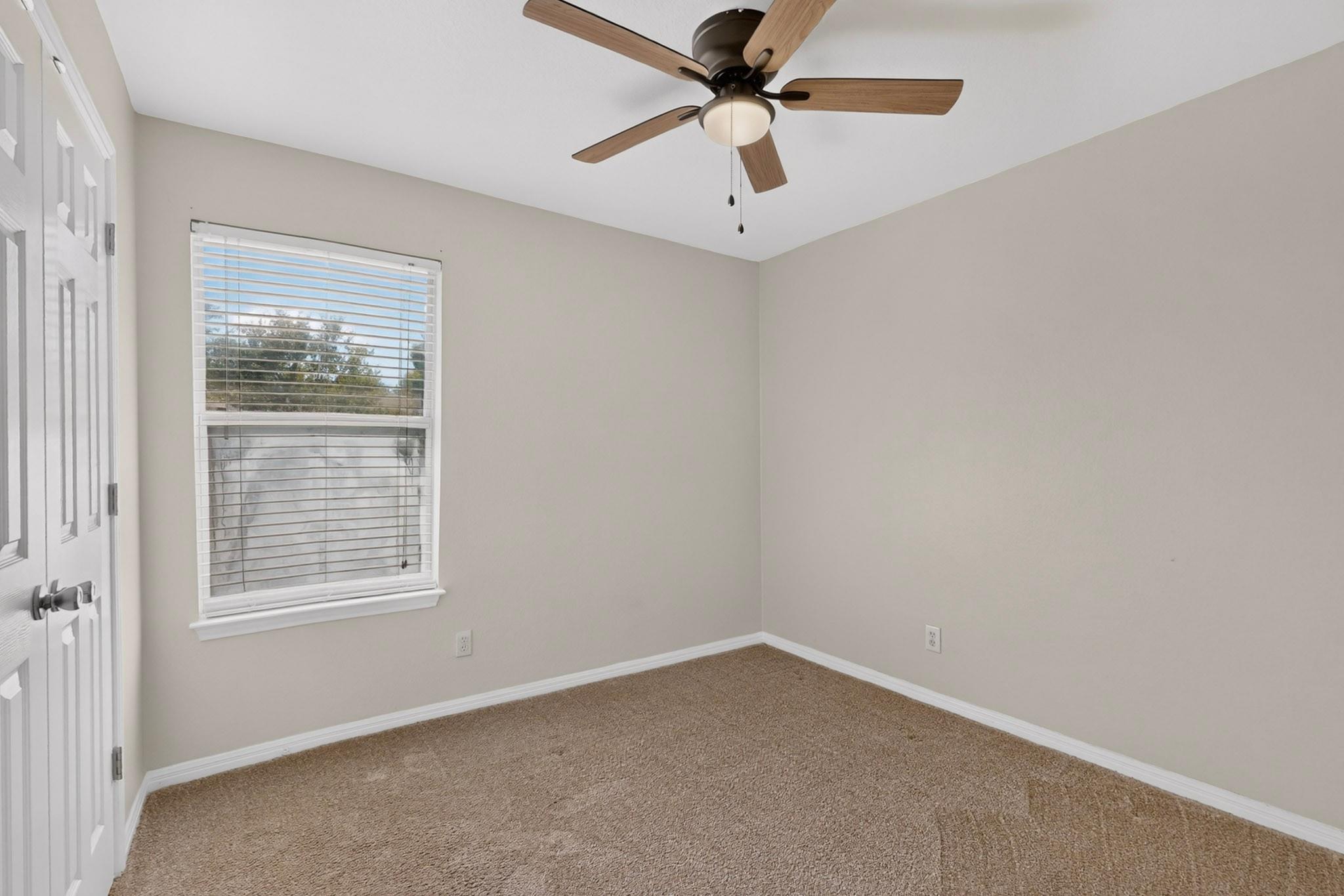 Empty carpeted bedroom with a window, white blinds, and a wood-blade ceiling fan.