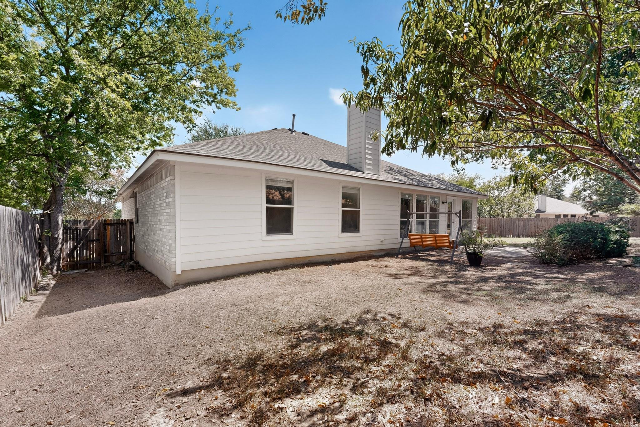 Rear of a white single-story house with a porch swing, mature trees, and a fenced yard.