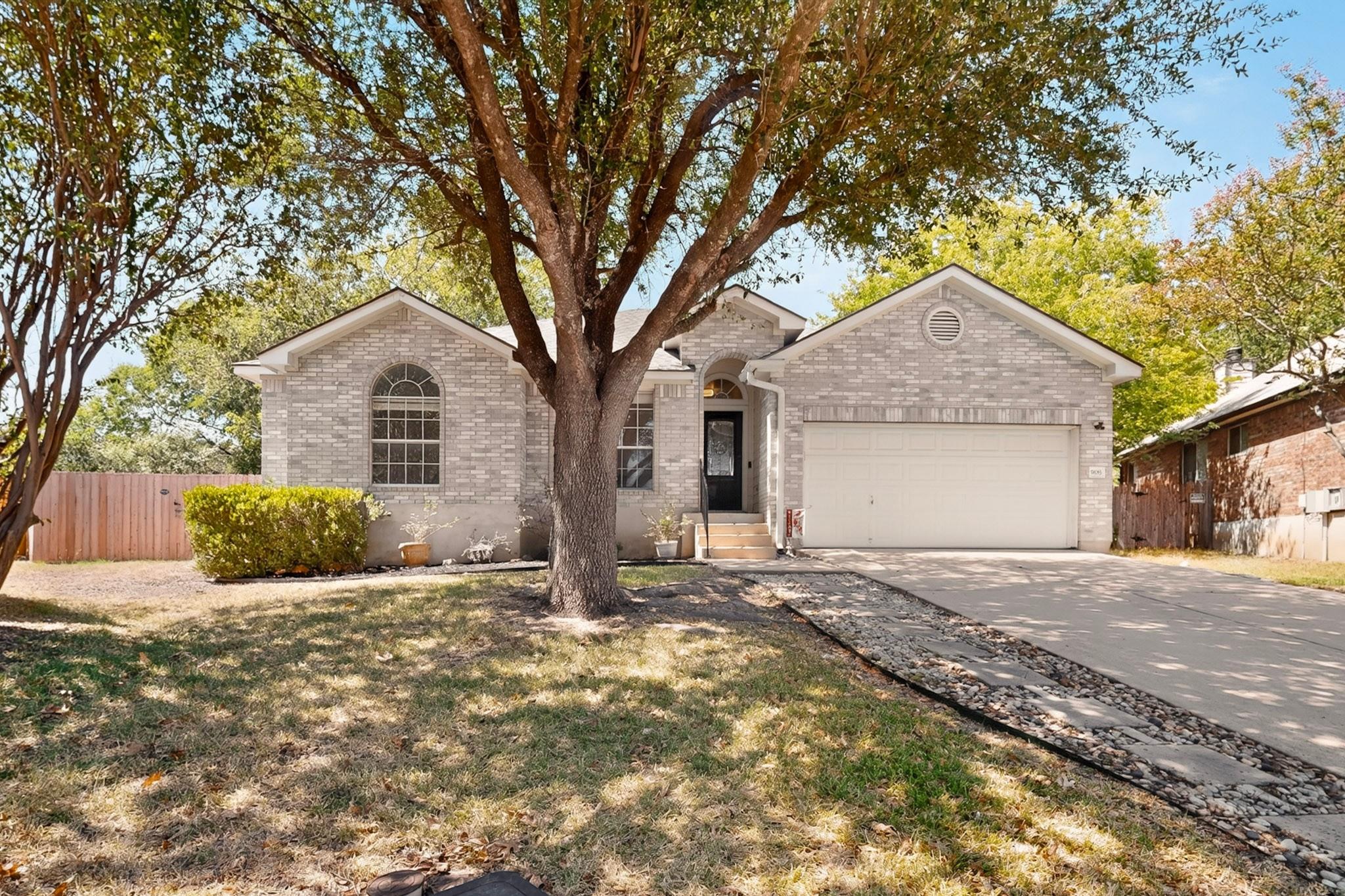 Light brick house with attached garage, arched windows, and a large tree in the front yard.