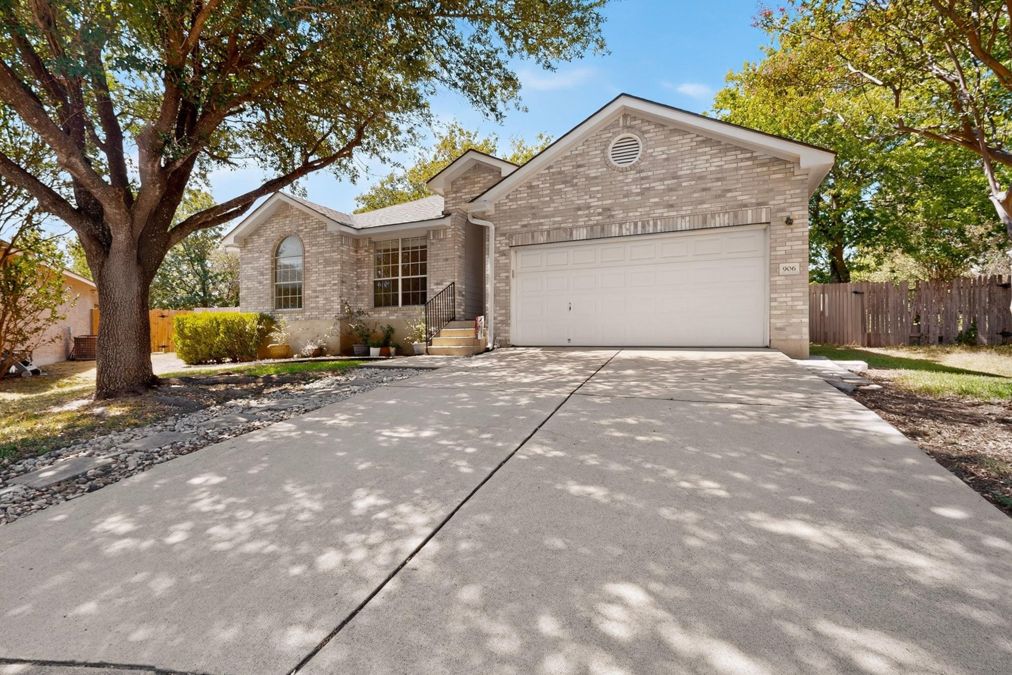 Light brick home with a two-car garage, broad driveway, and large shade tree.