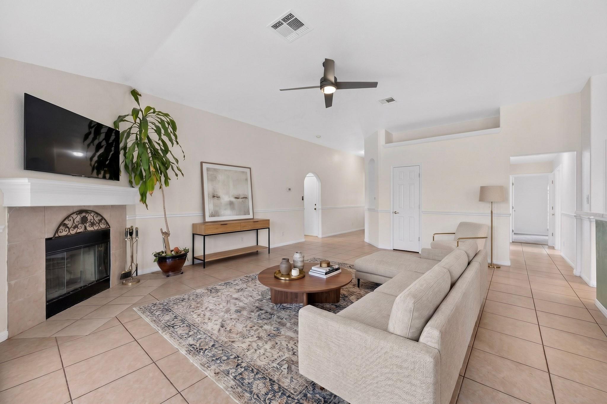 Staged living room with a beige sectional, tiled floor, and fireplace beneath a mounted television.