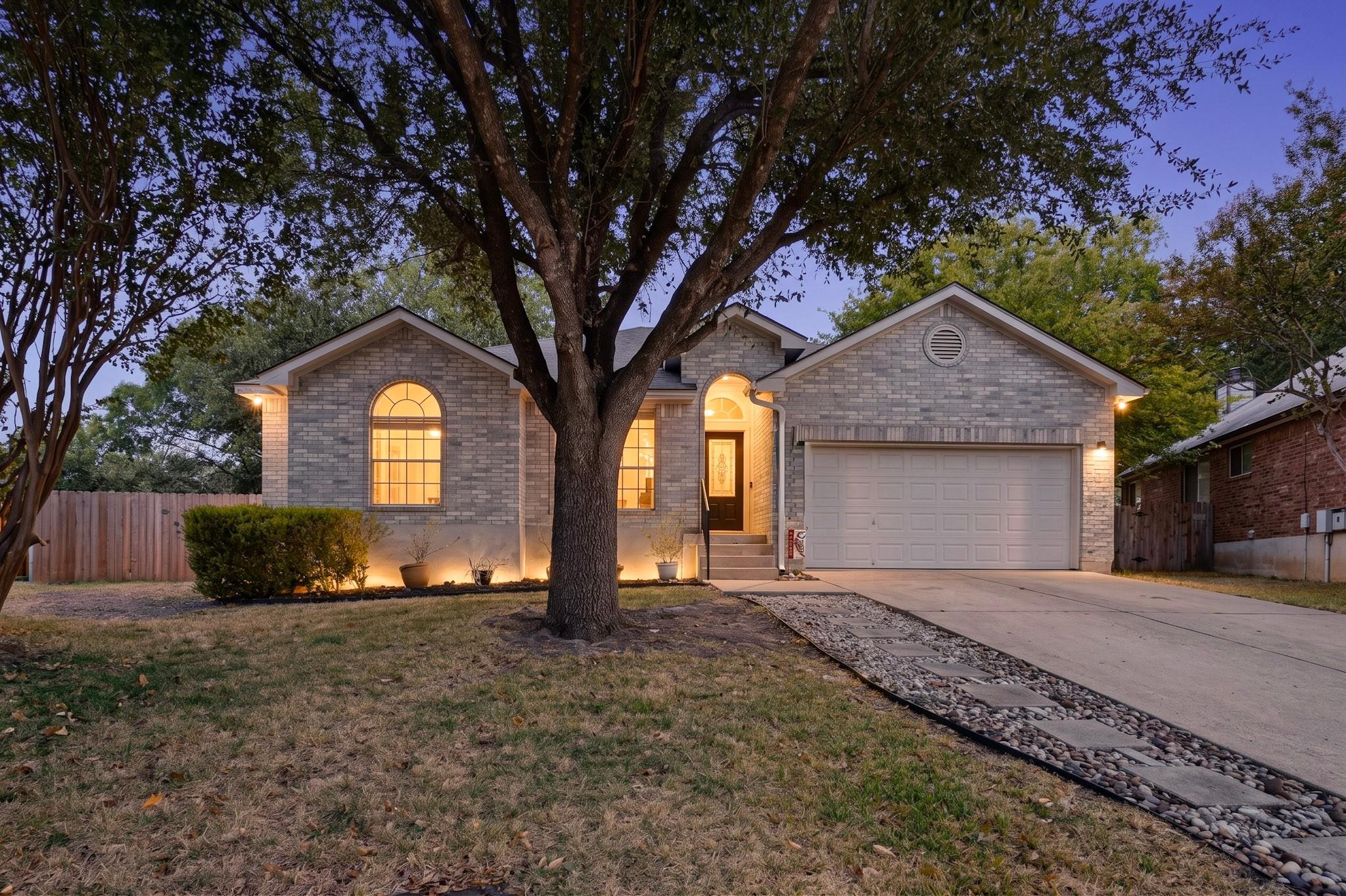 Brick home with warmly lit arched windows and entry, partly screened by a large front-yard tree.