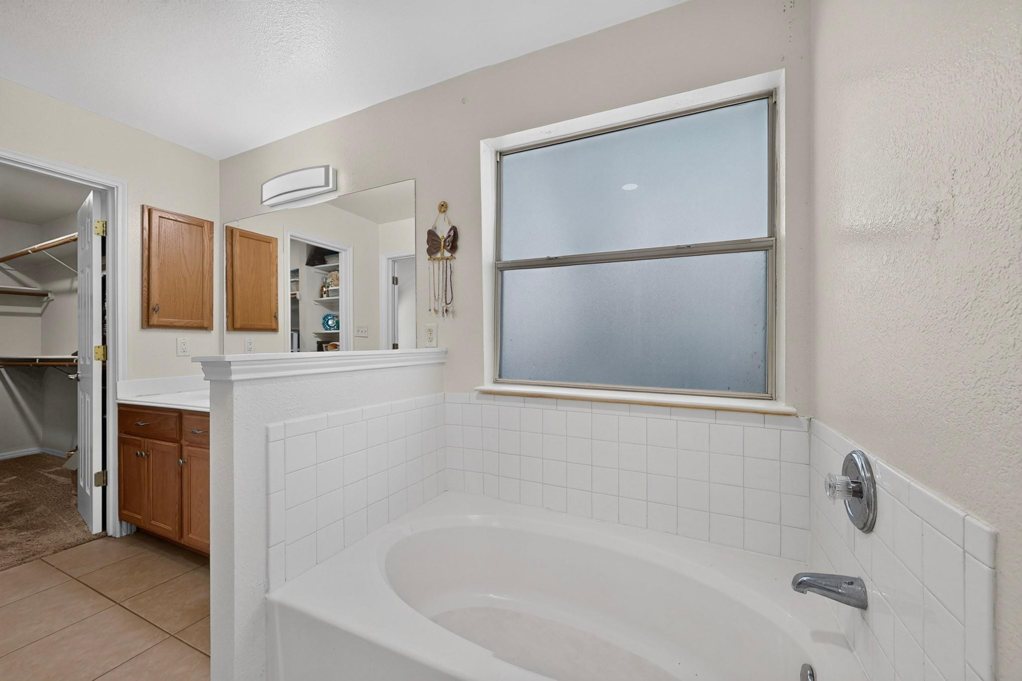 Bathroom with a large tiled bathtub beneath a frosted window, beside a wooden vanity and open closet.