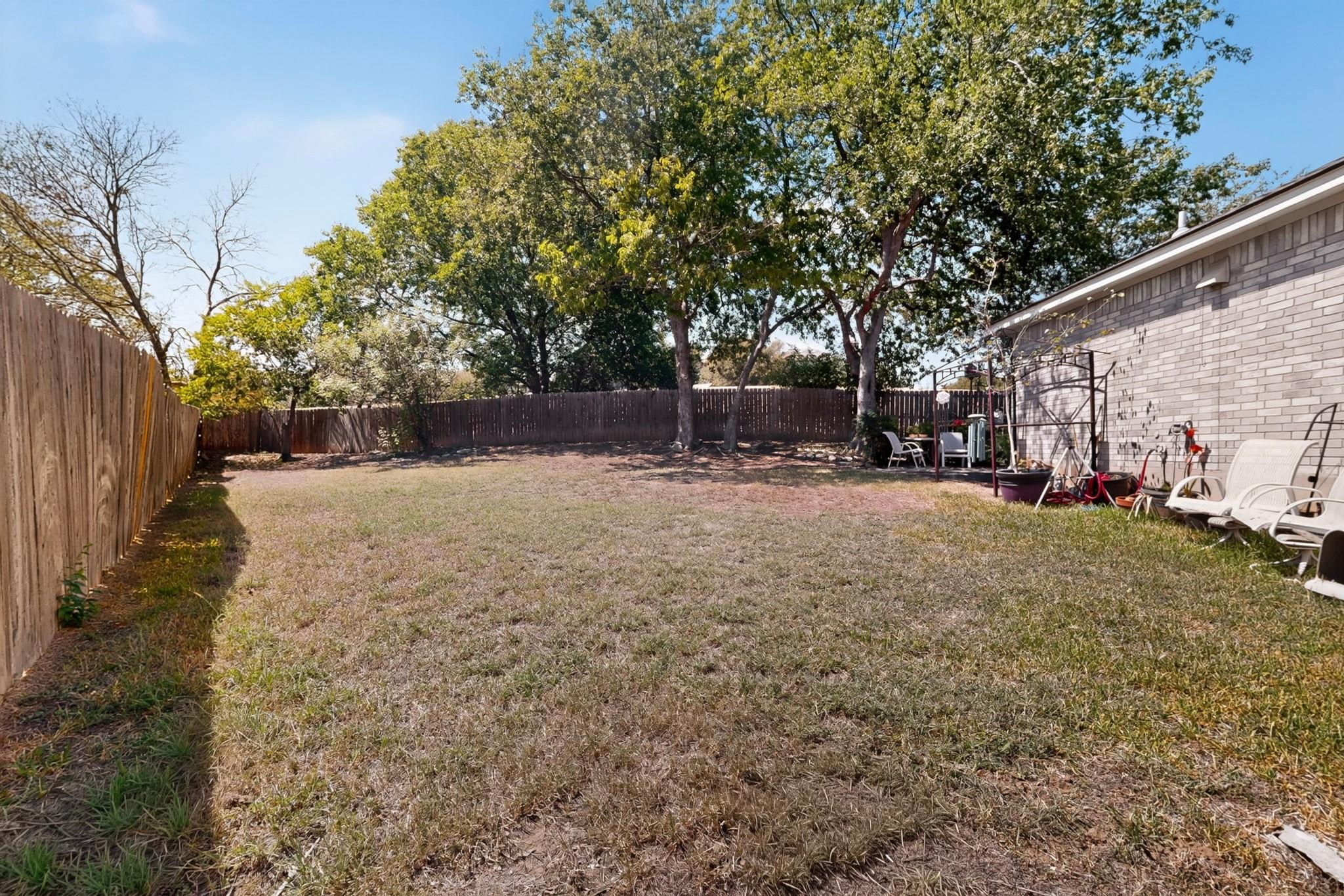 Fenced backyard with dry grass, mature shade trees, and chairs beside a brick house.