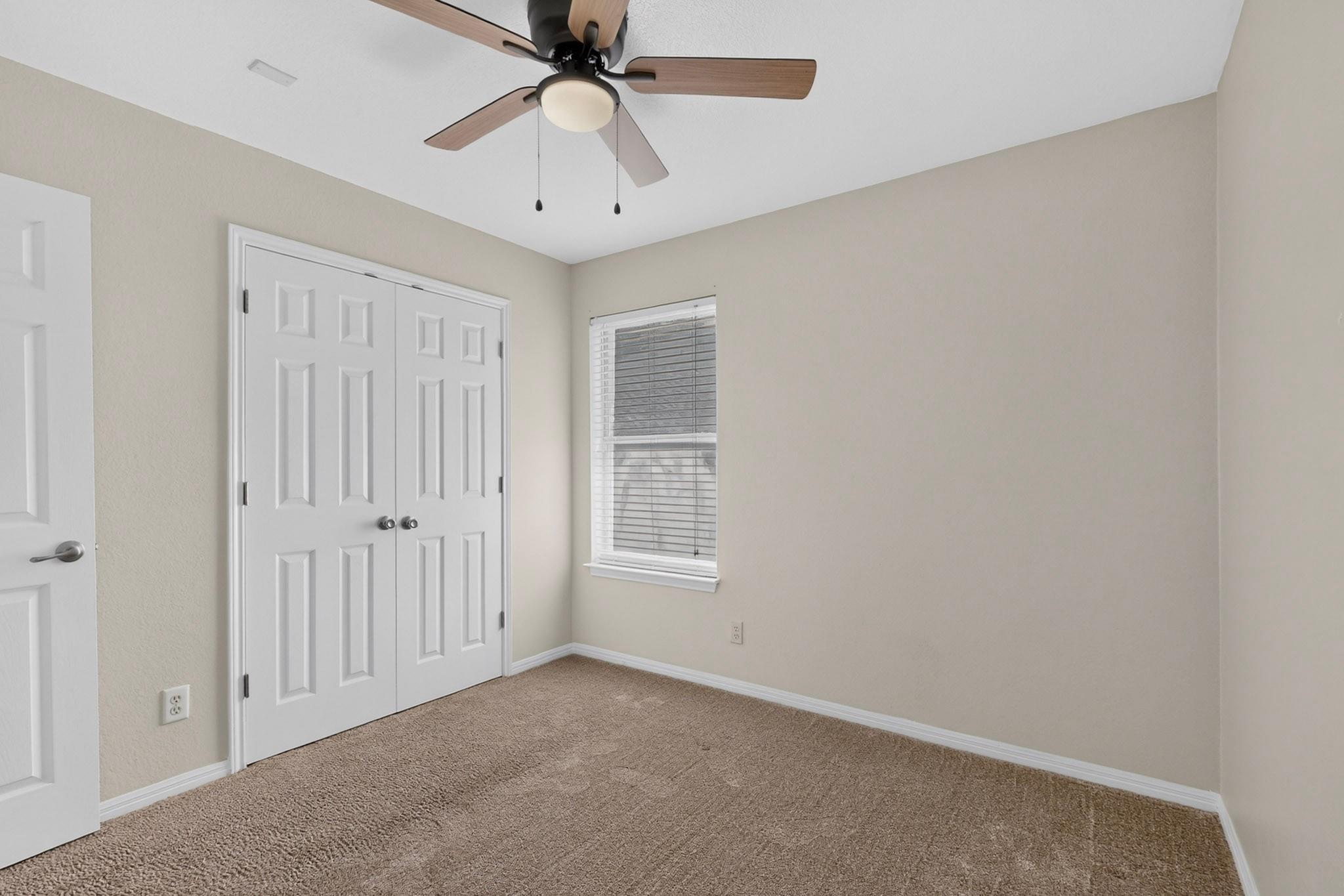 Unfurnished bedroom with beige carpet, double closet doors, a window with blinds, and a ceiling fan.