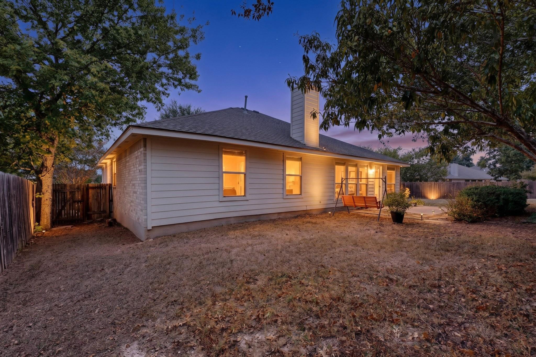 Rear of a single-story house glowing at dusk, with a porch swing and fenced, tree-shaded yard.