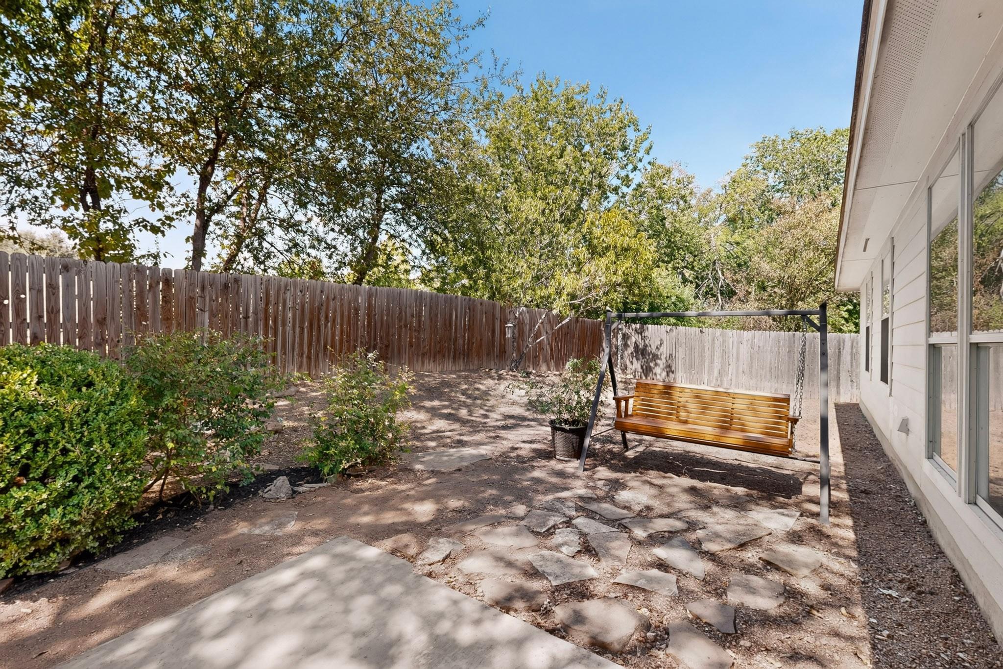 Backyard patio with a wooden swing, stone paving, mature trees, and a fenced yard.