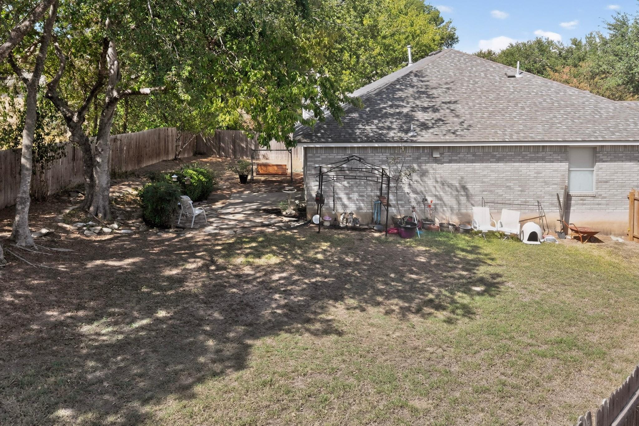 Fenced backyard beside a gray brick house, shaded by mature trees with patio chairs and a swing.