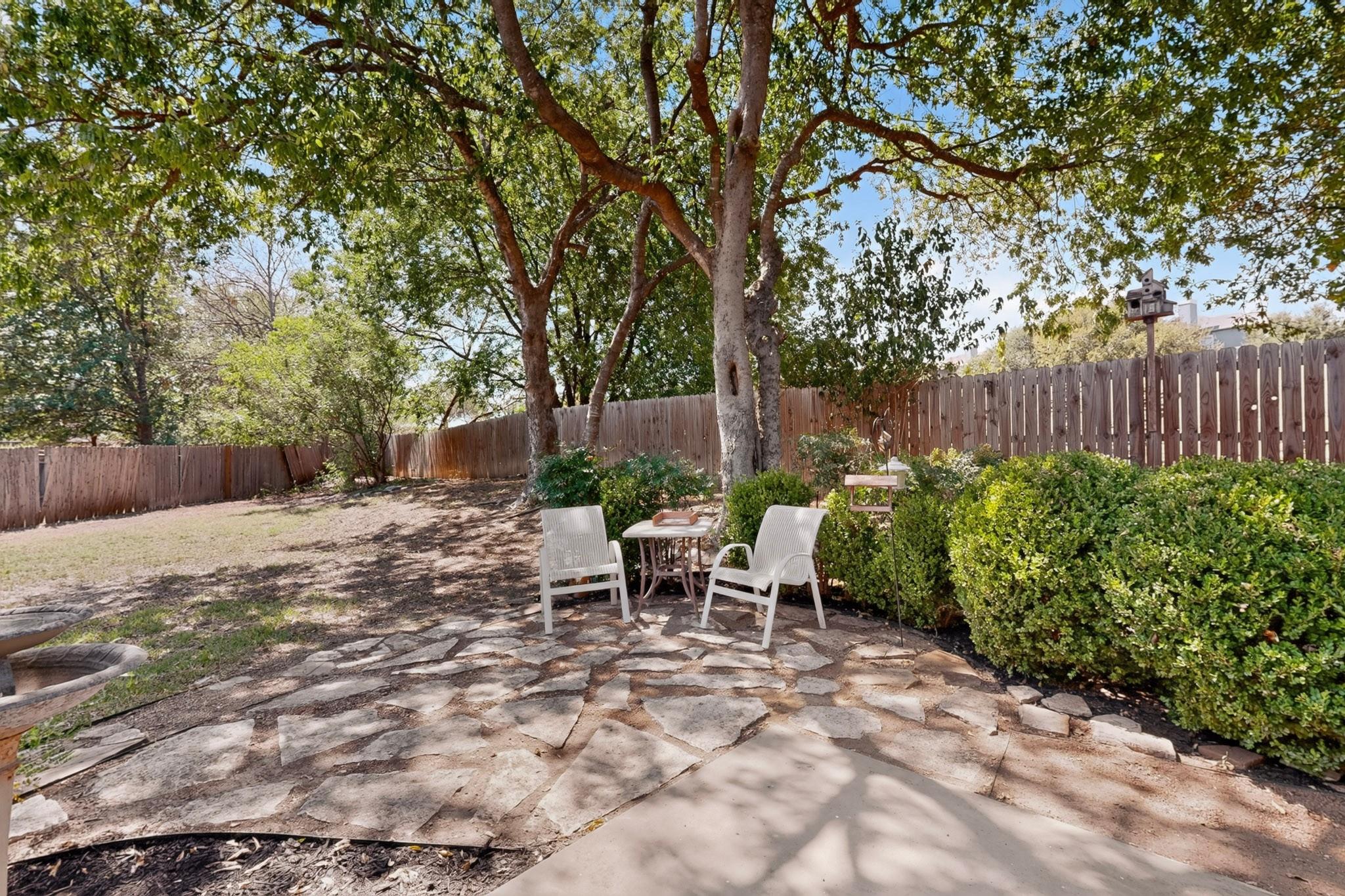 Shaded backyard patio with stone paving, white chairs, mature trees and a wooden fence.