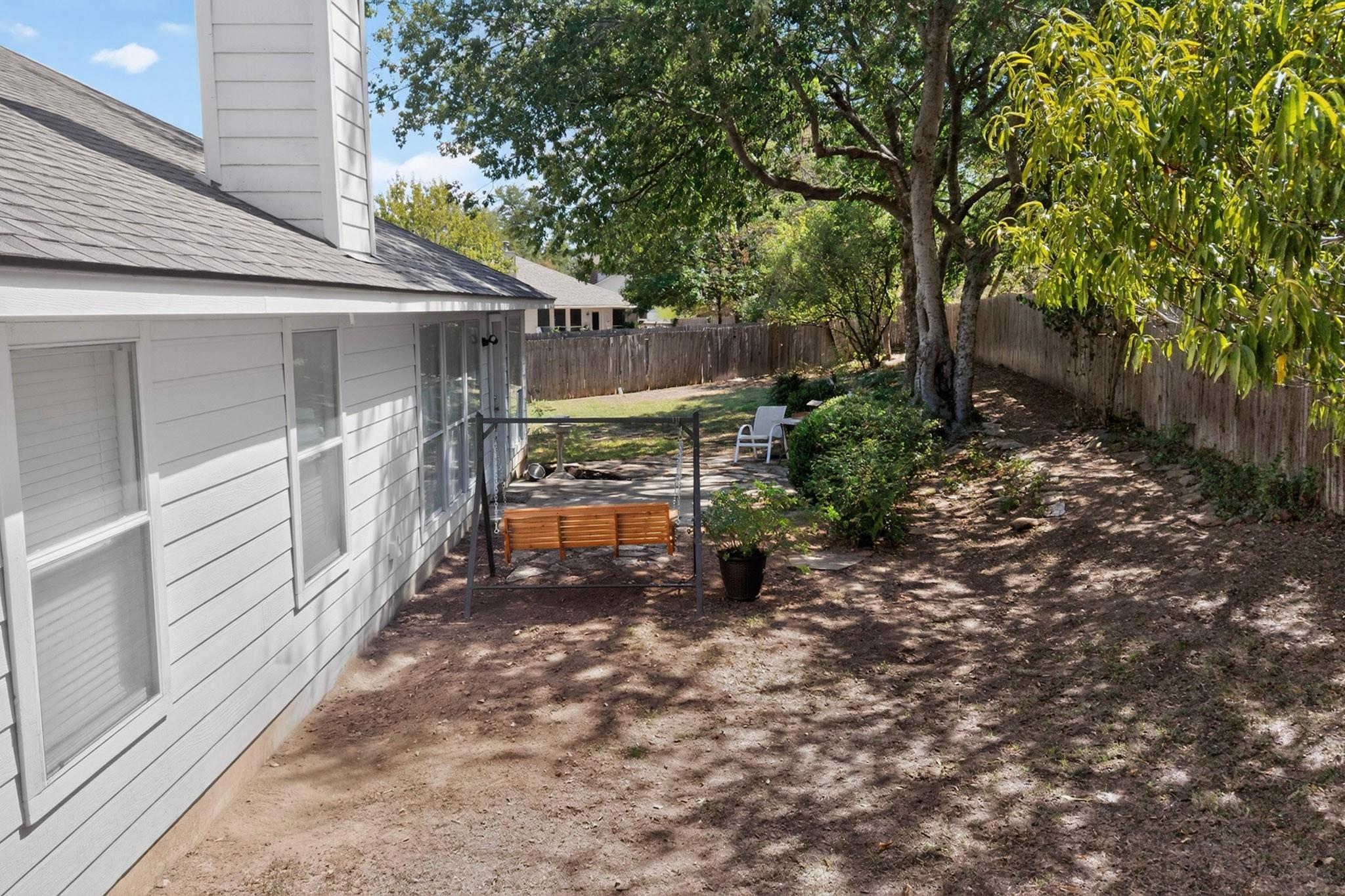 Fenced backyard with mature trees, a wooden swing beside the house, and outdoor seating.